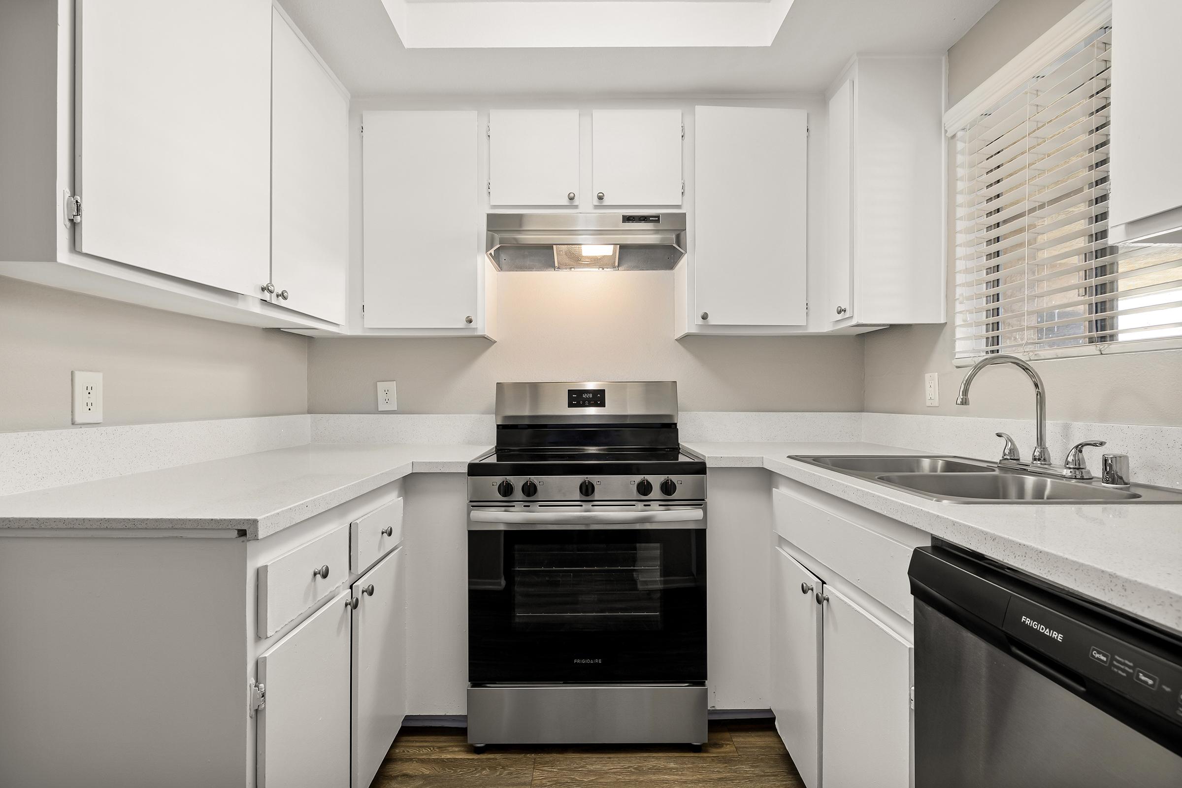 Modern kitchen featuring white cabinets, a stainless steel stove with an oven, a sleek range hood, and a double sink. The countertops are light-colored, and the space is illuminated by natural light from a window. The design is clean and contemporary, with a minimalistic aesthetic.
