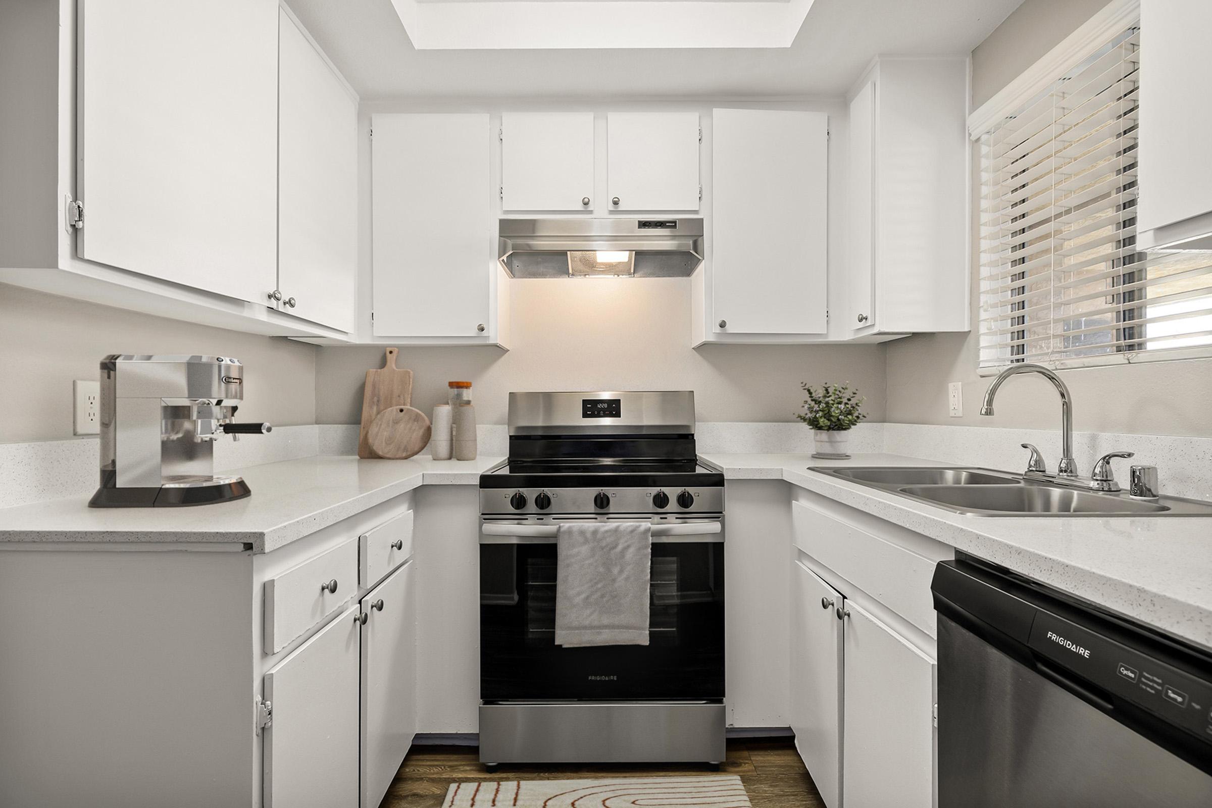A modern kitchen featuring white cabinets, a stainless steel stove with an overhead extractor, and a sink. A cutting board is placed on the counter, alongside kitchen utensils. Natural light from a window illuminates the space, complemented by a small potted plant. The floor has a decorative rug.