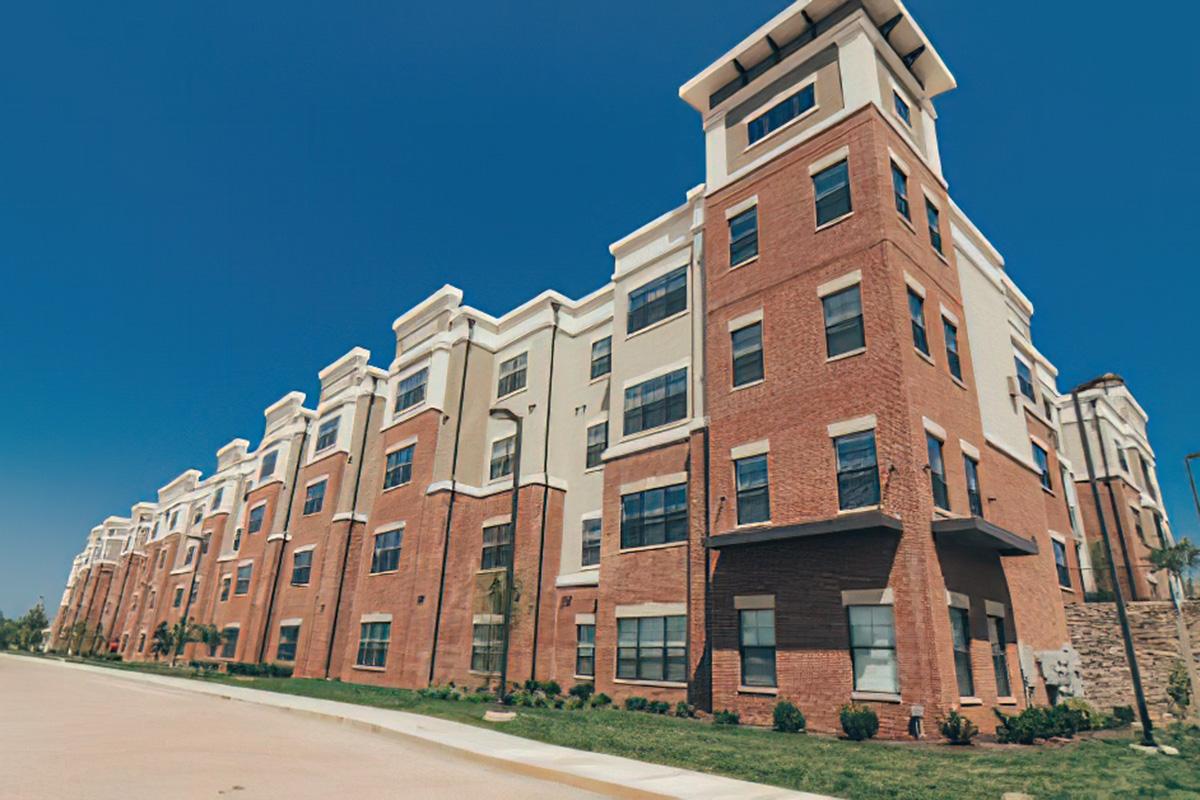 A modern brick apartment building with multiple stories and large windows, set against a clear blue sky. The structure features a variety of architectural details, including a peaked roof and balconies. Well-maintained landscaping is visible in the foreground, enhancing the aesthetic appeal of the building.