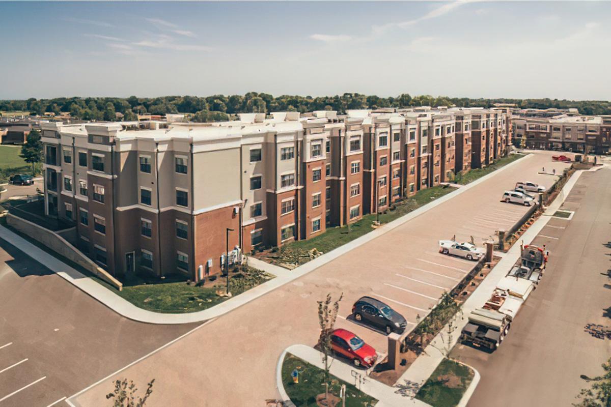Aerial view of a modern apartment complex featuring multiple three-story buildings with a mix of brick and siding. Surrounding landscaped areas include parking spaces, trees, and a wide driveway, showcasing a clean, organized residential community. Clear blue sky overhead.
