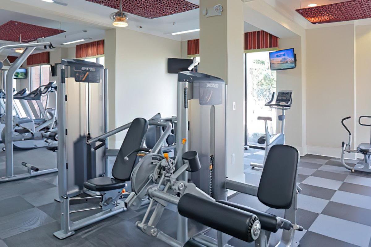 A well-equipped gym featuring various exercise machines including resistance and cardio equipment. Large windows provide natural light, and a television is mounted on the wall. The flooring is a mix of gray tiles, creating a modern and clean atmosphere.