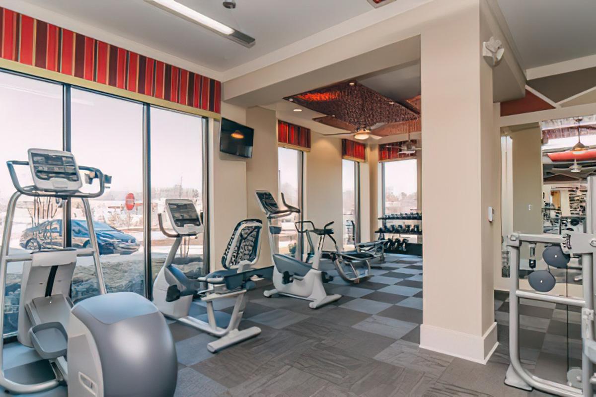 A modern gym interior featuring various fitness equipment, including treadmills, stationary bikes, and weights. Large windows allow natural light to fill the space, creating an inviting atmosphere. The area is well-organized with a contemporary design and decorative elements on the ceiling.