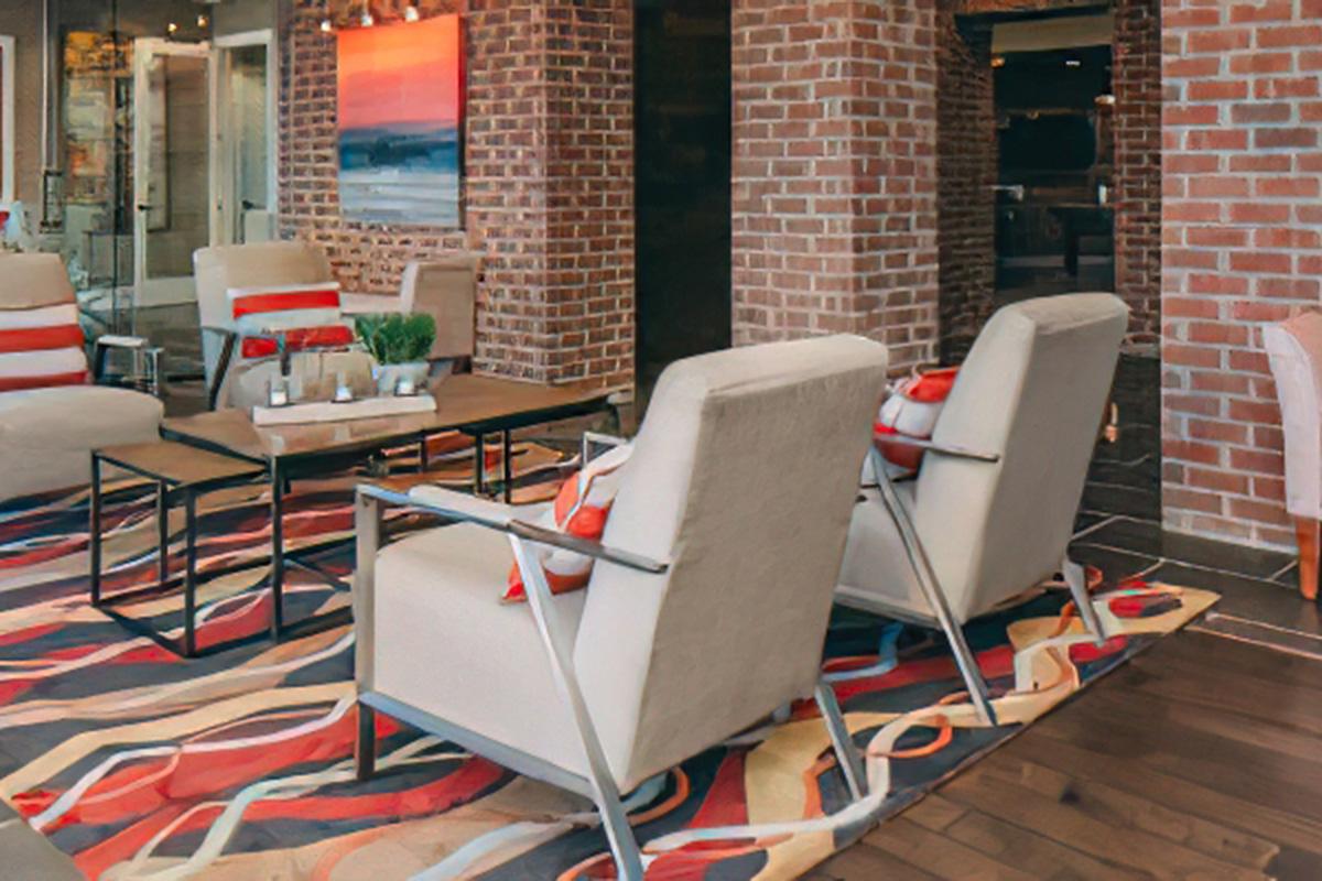 A modern living space featuring two light gray armchairs with red cushions, a dark coffee table, and a colorful area rug. The room has exposed brick walls, large windows, and contemporary artwork on display, creating a stylish and inviting atmosphere.