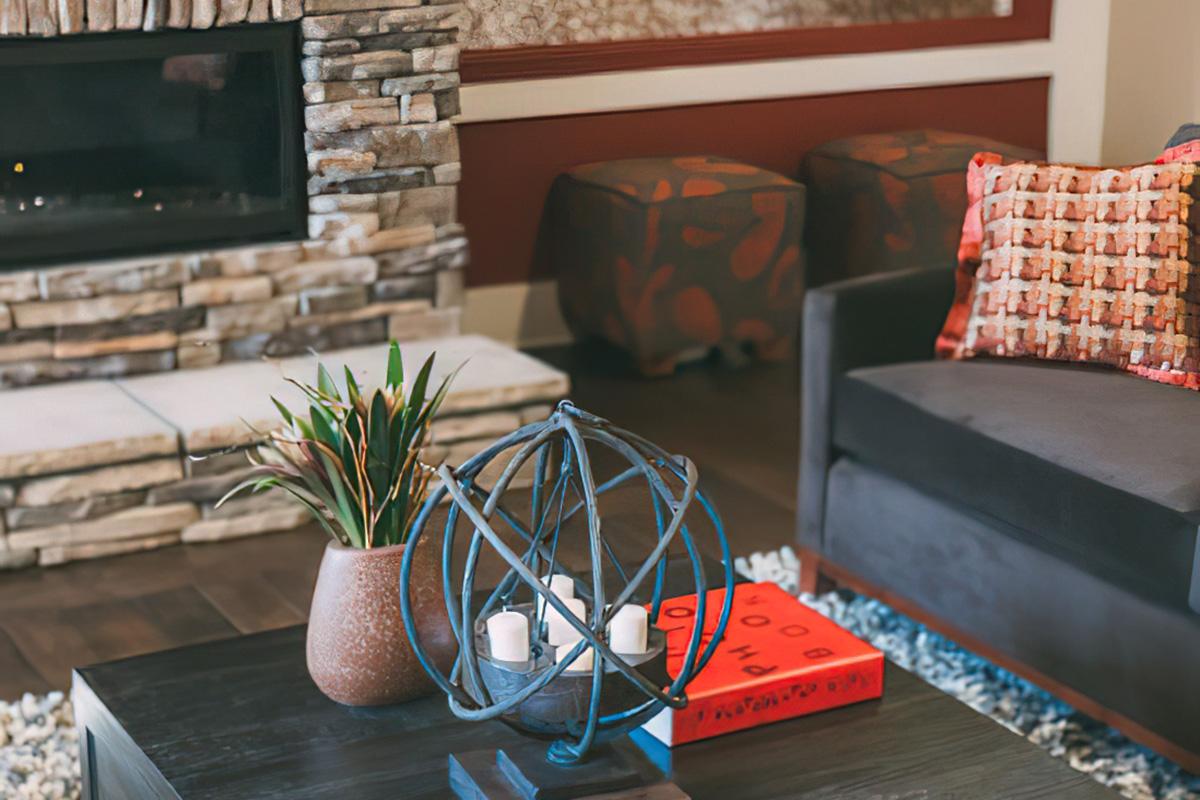 A cozy living room featuring a stone fireplace, a dark sofa with a decorative pillow, a wooden coffee table with a decorative orb and candles, and a potted plant. Two colorful ottomans are visible in the background, adding to the inviting atmosphere.