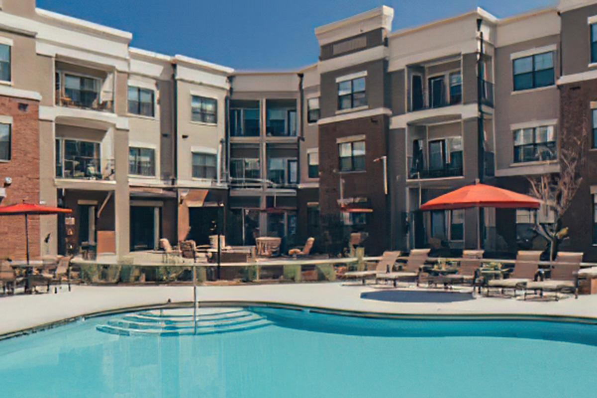 A sunny view of an apartment complex featuring a swimming pool in the foreground. The pool is surrounded by lounge chairs, and there are several orange umbrellas providing shade. The buildings in the background are modern with balconies and large windows, creating a welcoming outdoor space.