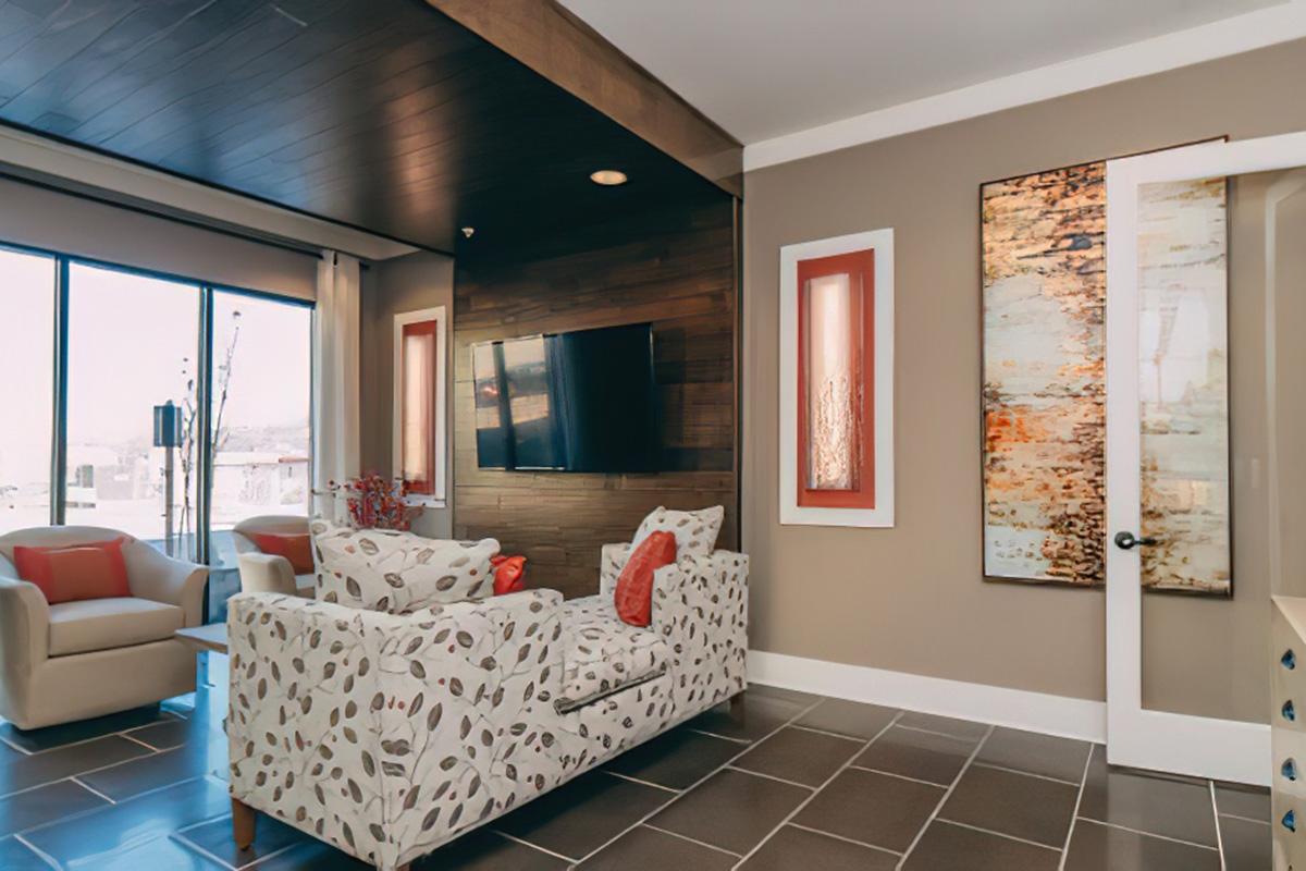 A modern living room featuring a floral-patterned couch, two orange accent chairs, and a wall-mounted TV. Natural light streams in through large windows, illuminating the space. A piece of abstract artwork is displayed on the wall, complementing the neutral color scheme and contemporary design elements.
