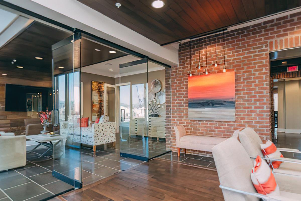 Modern office interior featuring a glass partition with a view to an adjoining space. The room has a warm color palette with brick walls, wood accents, and stylish seating. A contemporary painting hangs on the wall, while the floor combines tiles and wooden elements, creating a welcoming atmosphere.