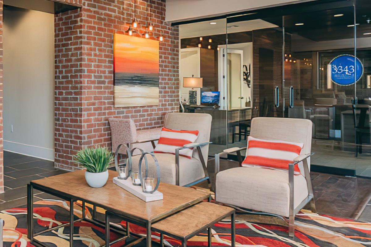 A cozy interior space featuring two light-colored armchairs with orange striped cushions, a wooden coffee table with decorative items, and a vibrant abstract painting on a brick wall. The area has modern lighting and a glass partition showcasing a contemporary office space.
