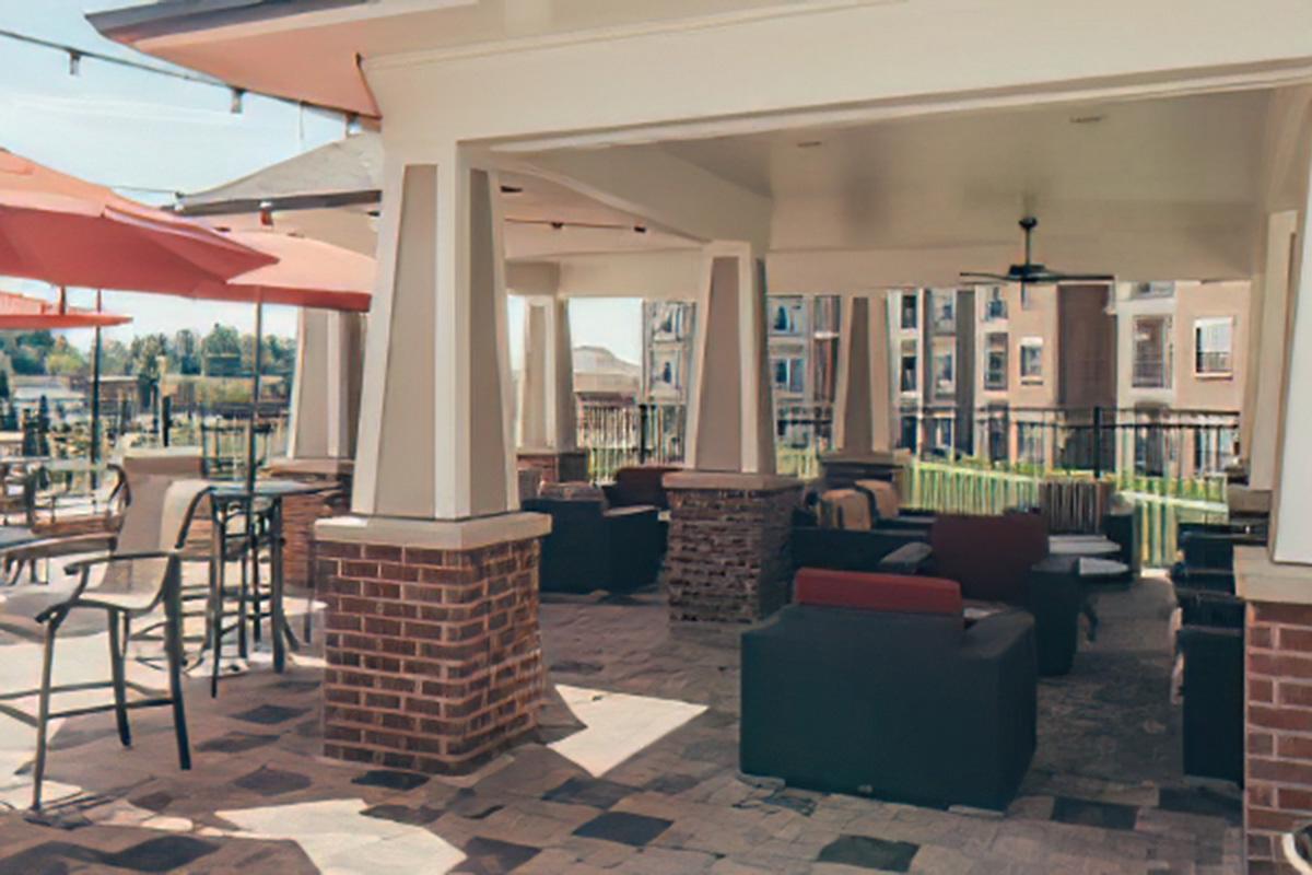 Outdoor lounge area featuring comfortable seating with dark and light cushions, tall tables, and red umbrellas. The space is surrounded by green landscaping and has a view of nearby buildings, creating a relaxing atmosphere for social gatherings.
