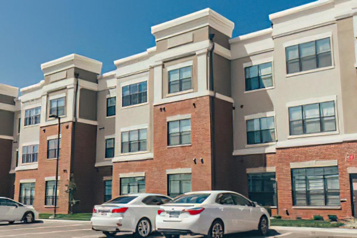 A modern multi-story apartment building with a mix of brick and painted surfaces. The facade features large windows and decorative cornices. Two parked white cars are visible in front, and the sky is clear and blue, providing a bright setting.
