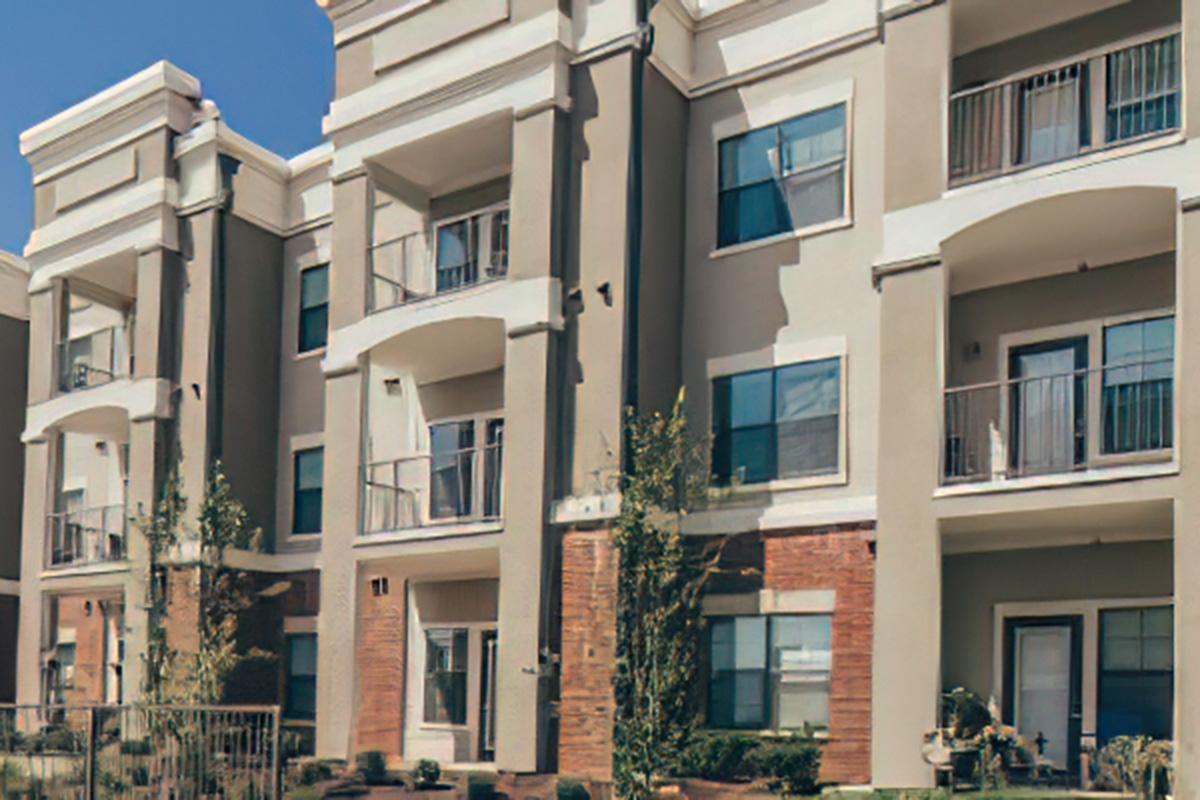 A modern, multi-story residential building with a beige facade, large windows, and balconies. The structure features a mix of materials, including brick accents and greenery around the entrance, set against a clear blue sky.