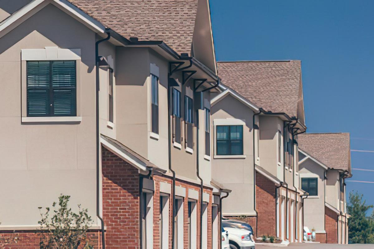 Row of modern residential buildings featuring a mix of brick and smooth exterior walls, with multiple windows and gabled roofs under a clear blue sky. The foreground shows a driveway with parked cars and well-maintained landscaping.