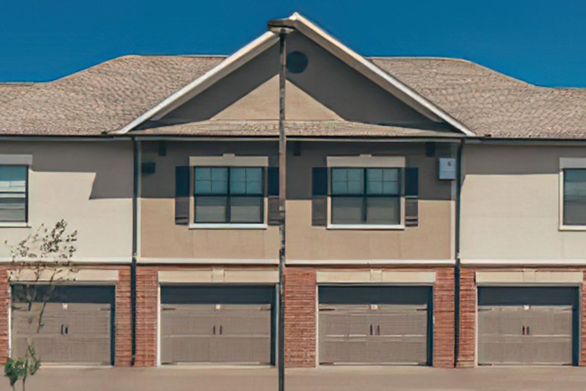A row of cream-colored townhouses with brown brick accents and garage doors. The buildings feature a gabled roof and symmetrical window placement, with large windows framed by shutters. The clear blue sky creates a bright backdrop for the homes.