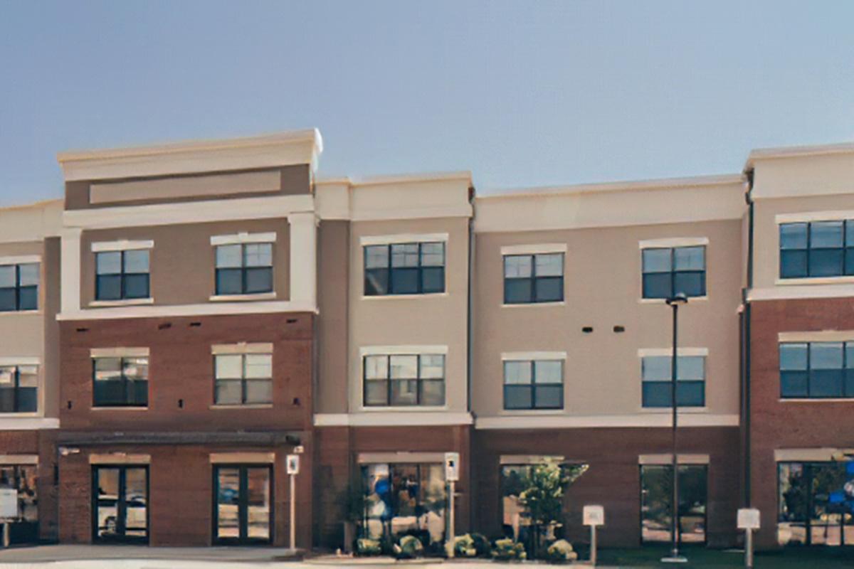 Modern multi-story building with a mix of brown and beige exterior, featuring large windows and a flat roof. The ground floor has a storefront area, and there are signs and greenery in the vicinity. Clear blue sky above.