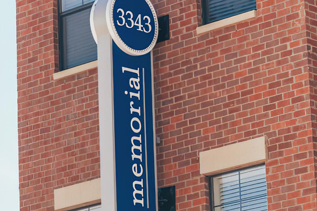 Sign on a brick building featuring the number "3343" and the word "memorial" in white text on a dark blue background. The sign is prominently displayed, indicating a specific location or establishment. Windows of the building are visible in the background.