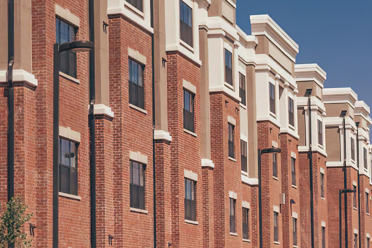 A row of modern brick apartment buildings with large windows and decorative trim against a clear blue sky. The architecture features a contemporary design, showcasing a combination of red brick and light-colored accents.