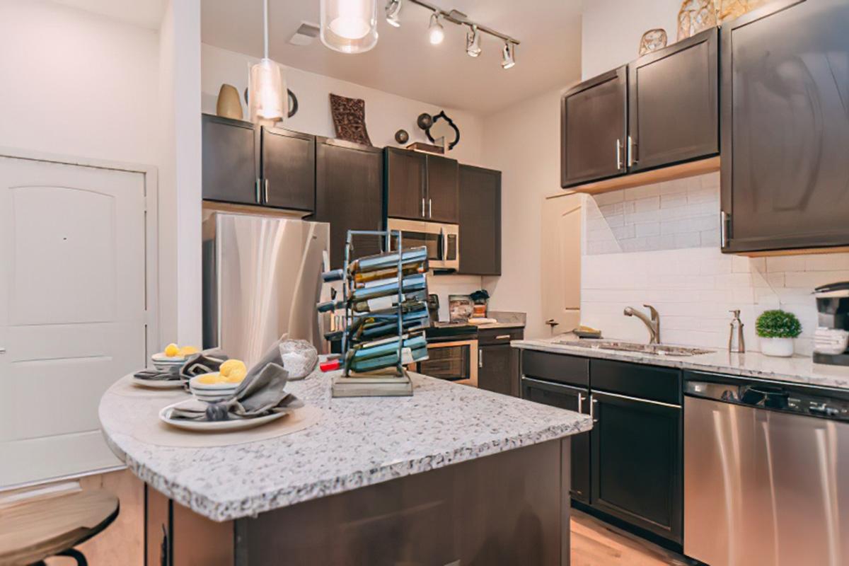 Modern kitchen featuring dark cabinetry, stainless steel appliances, and a granite countertop island. The island is set with plates, napkins, and decorative items, while pendant lighting illuminates the space. A door is visible in the background, adding to the inviting atmosphere.