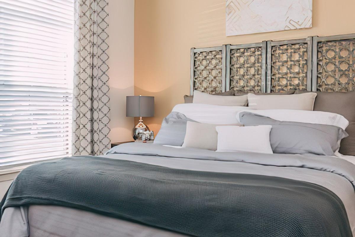 Cozy bedroom featuring a large bed with various gray and white pillows, adorned with a textured throw blanket. A decorative headboard adds elegance, while a bedside table holds a stylish lamp. Light filters through window blinds, creating a warm and inviting atmosphere.