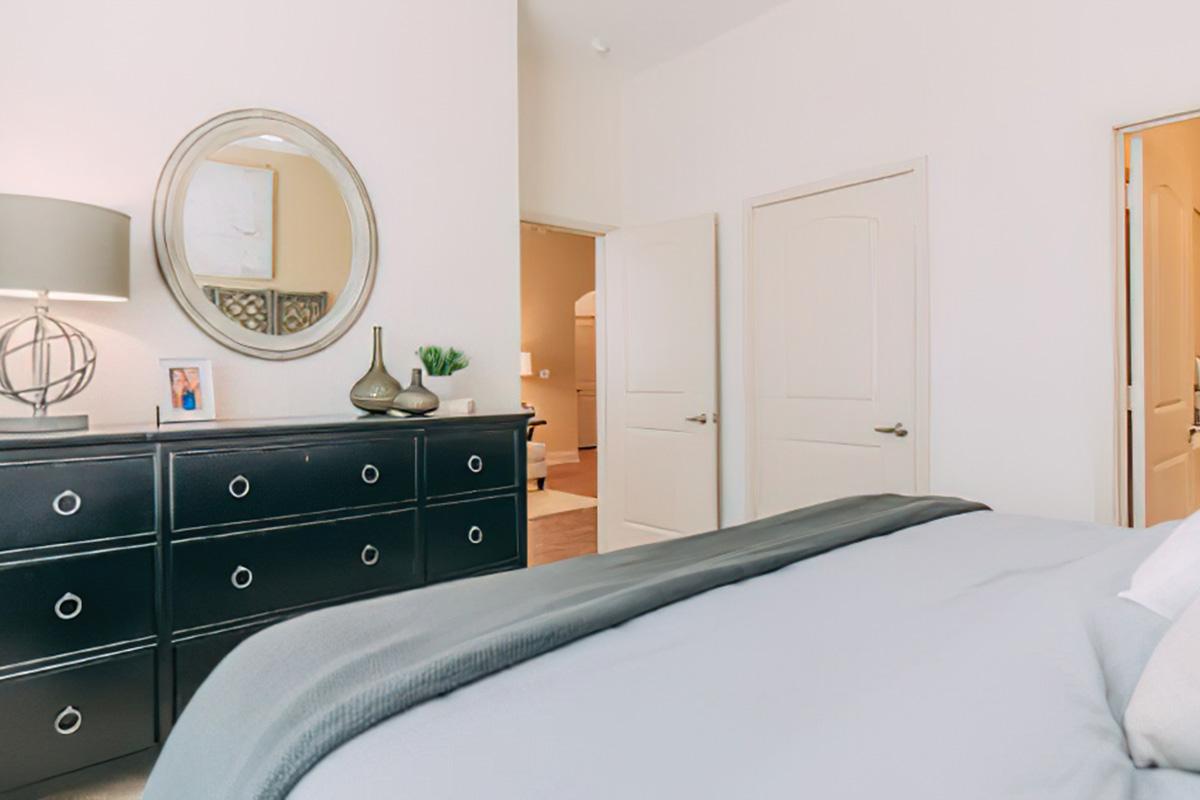 A well-decorated bedroom featuring a dark dresser with decorative items, a round mirror on the wall, and a neatly made bed with gray bedding. Two doors are visible in the background, leading to other rooms. The overall aesthetic is modern and inviting.