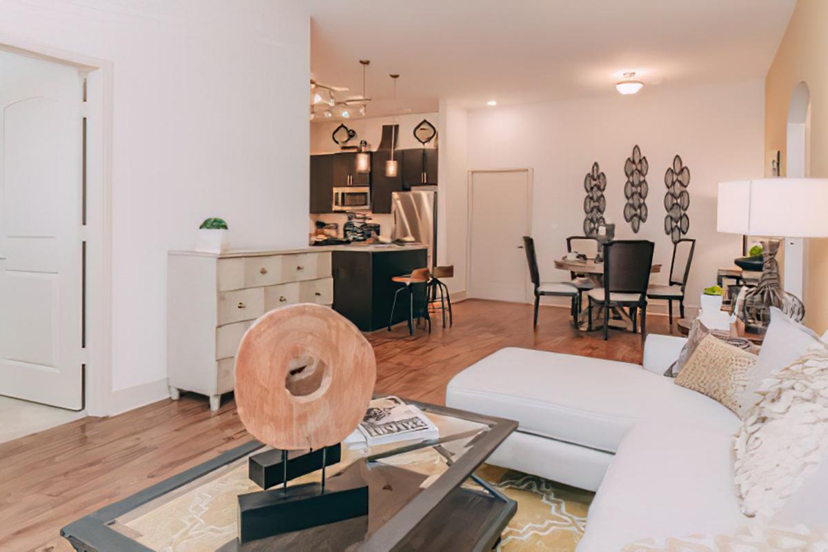 A modern living room featuring a white sectional sofa, a wooden accent table with a circular sculpture, and a coffee table with magazines. The background shows a dining area with black chairs, a kitchen with dark cabinetry, and decorative wall art. The space is well-lit and welcoming.