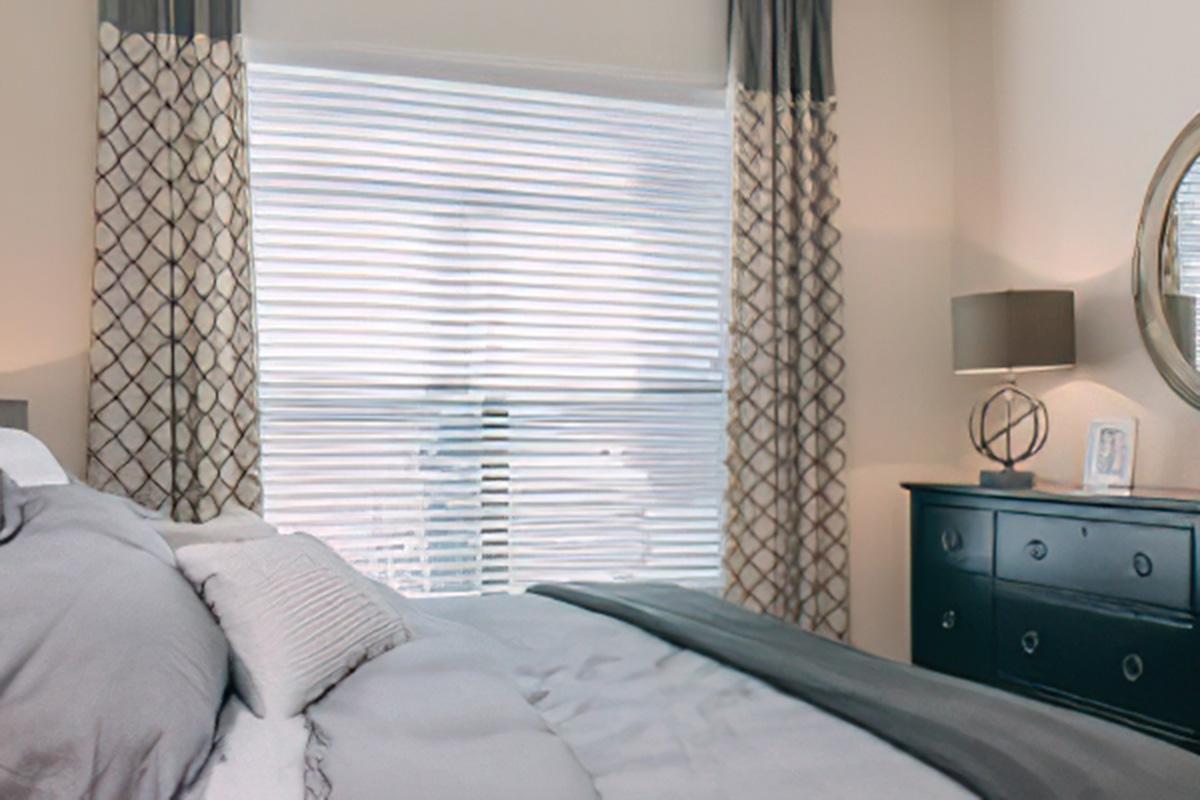 A stylish bedroom featuring a neatly made bed with gray bedding, decorative pillows, and a patterned curtain over a large window. A round mirror hangs on the wall above a dark wooden dresser, which has a decorative accent. Natural light filters through the window, creating a serene atmosphere.