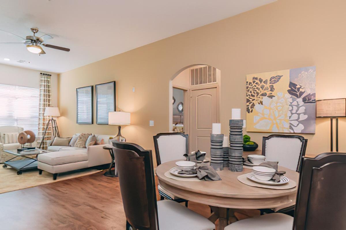 Modern living space featuring a dining table set with plates and candles, a cozy sofa with decorative pillows, and an artwork on the wall. Natural light filters through window treatments, and there are neutral tones with touches of greenery. The decor is inviting and stylish.