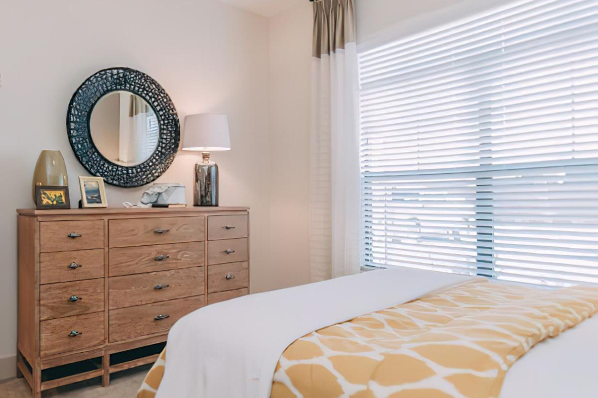 A cozy bedroom featuring a light-colored wall and large window with blinds. There's a wooden dresser with nine drawers, a round mirror above it, and a bedside lamp. A yellow and white patterned throw blanket is laid over the bed, adding a vibrant touch to the serene space.