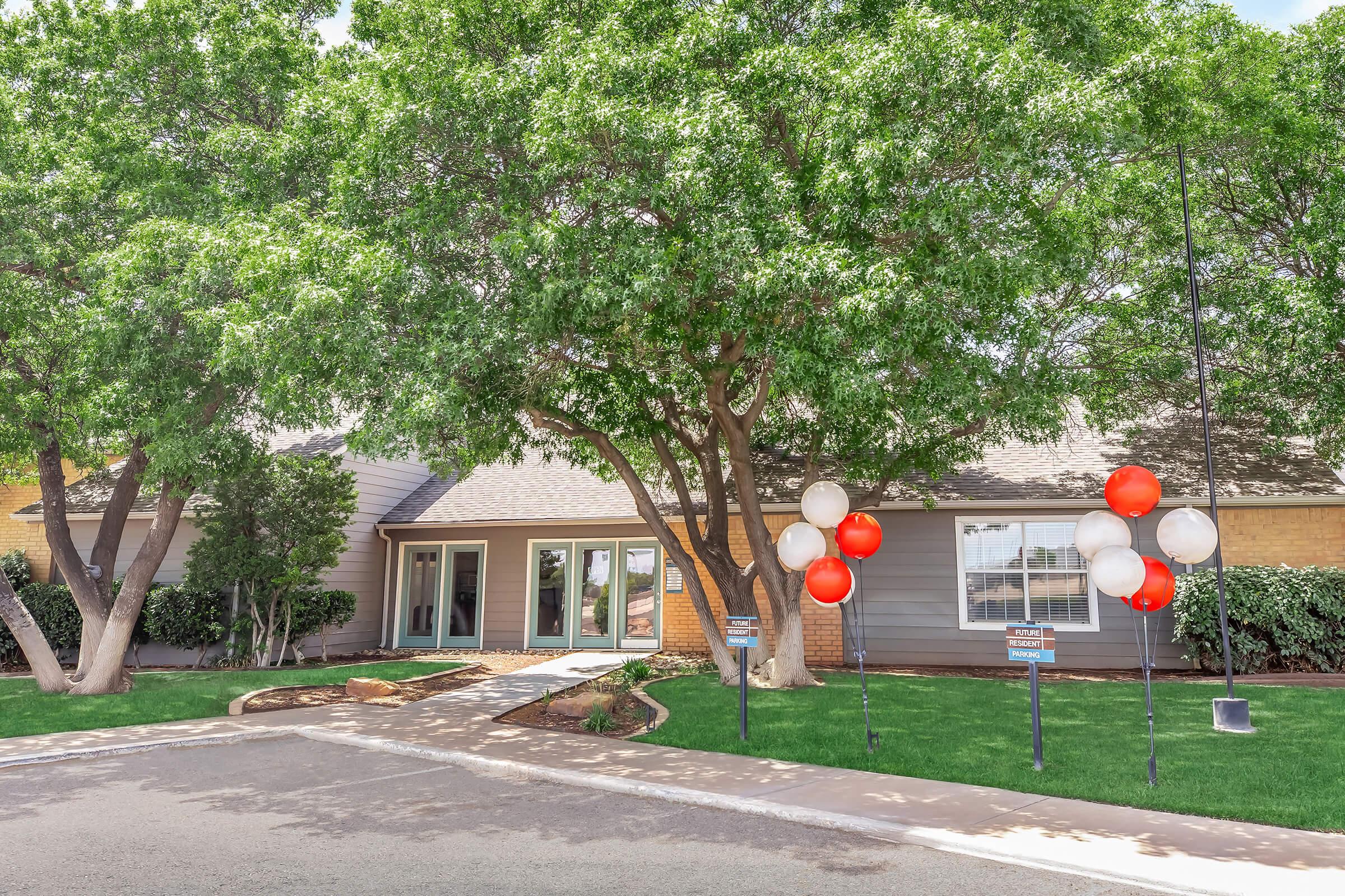 A modern single-story house with a light gray exterior, large windows, and a welcoming entryway is surrounded by green trees. Red and white balloons are tied to signposts in the foreground, adding a festive touch to the well-maintained front yard and walkway.