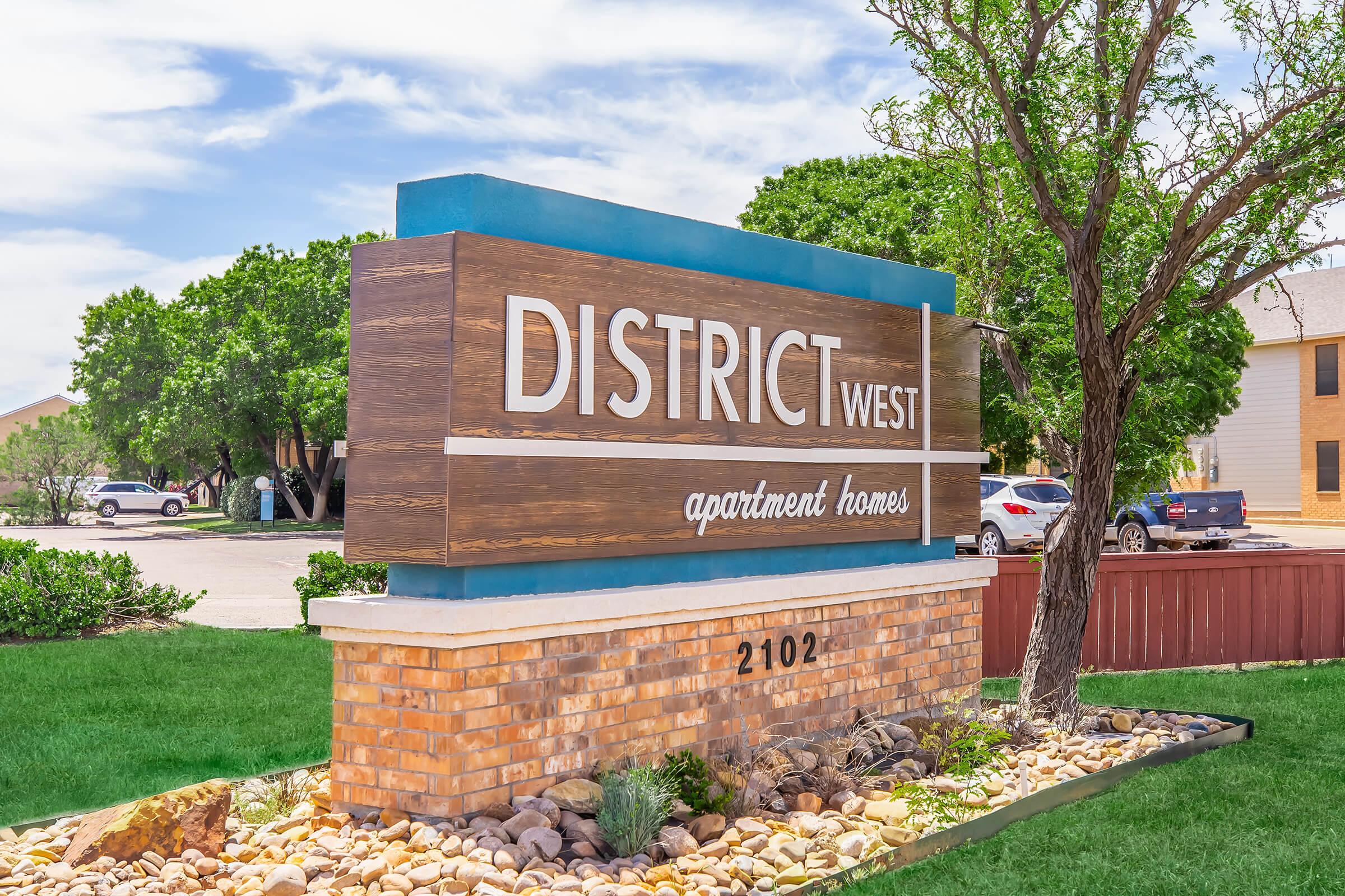 Sign for District West apartment homes featuring a wooden and blue design on a brick pedestal, surrounded by grass and decorative stones. In the background, there are trees and parked cars, indicating a residential area. The address "2102" is displayed at the bottom of the sign.