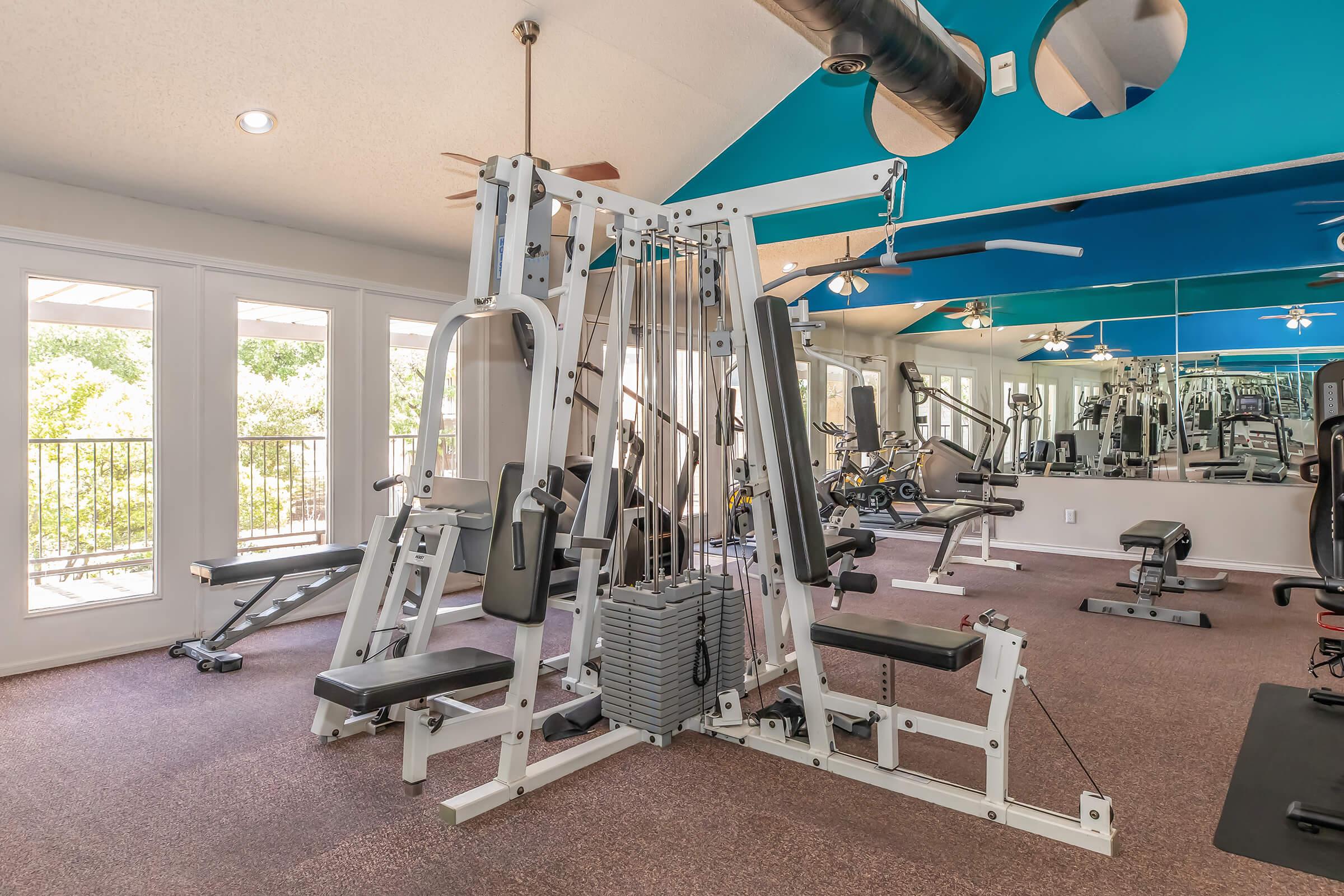 A modern gym interior featuring a multi-station weight machine at the center, surrounded by various exercise equipment. The space has large windows allowing natural light, with blue ceiling accents and ceiling fans for ventilation. The flooring is carpeted, creating a comfortable workout environment.