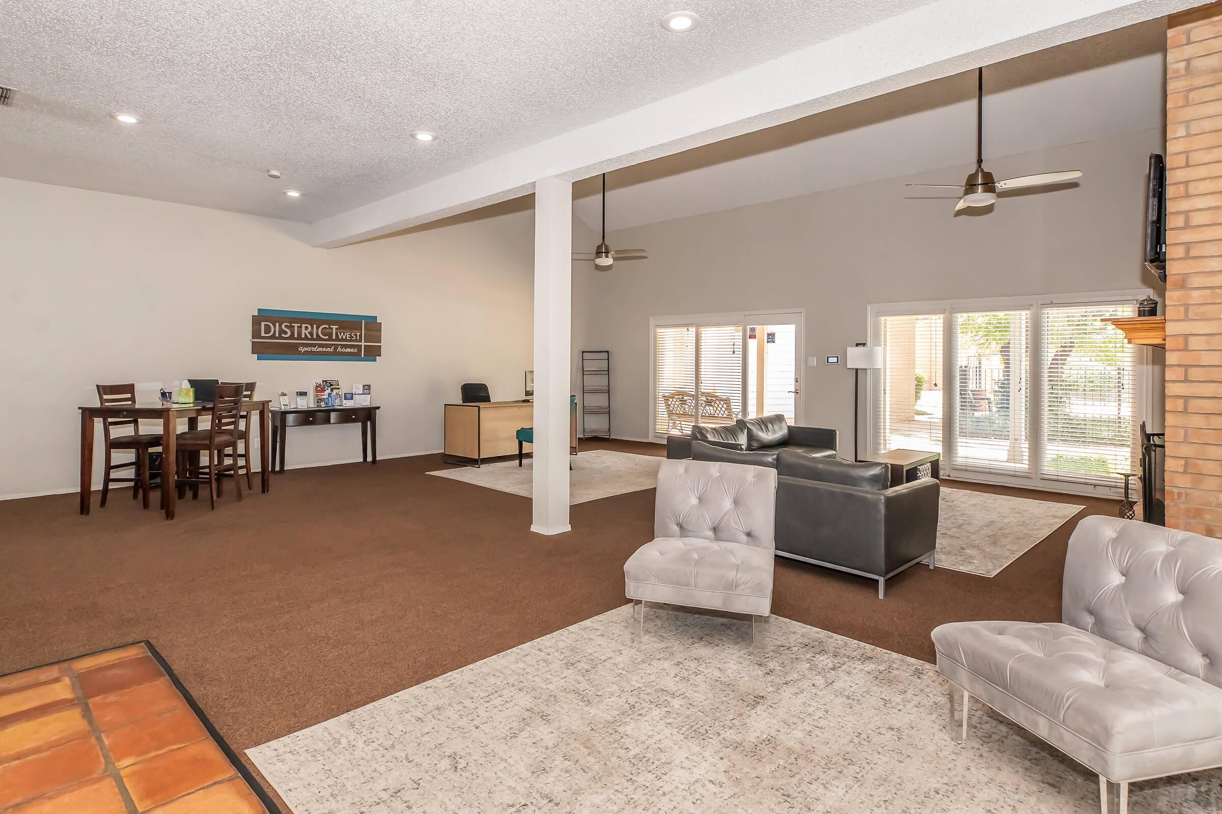 A spacious, modern living area featuring light-colored walls and a high ceiling. There are two comfortable seating areas: one with a gray sofa and a chair, and another with a dining table and four chairs. Large windows let in natural light, and there's a small desk area visible. The floor is carpeted and a patterned rug adds warmth.