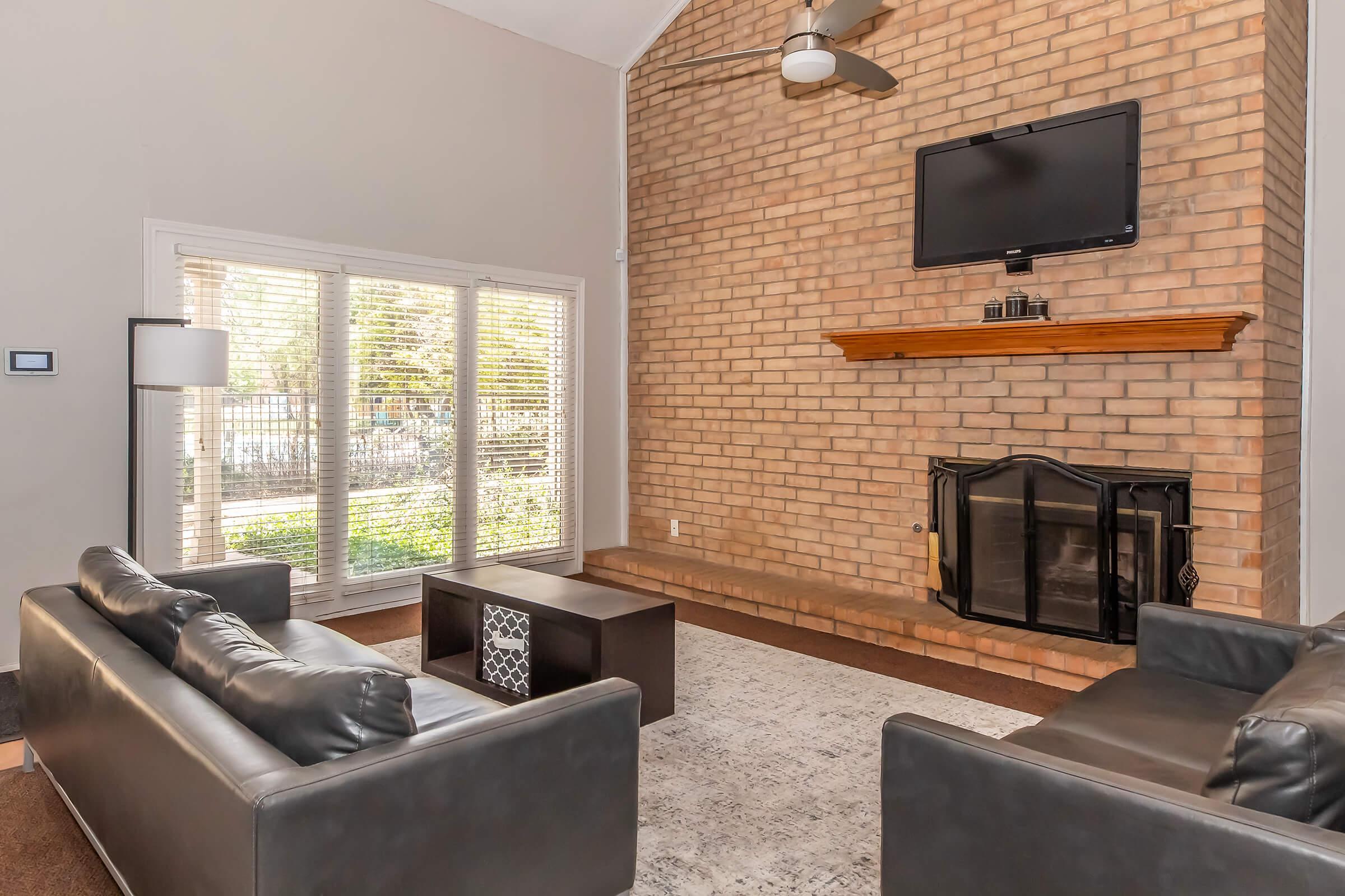 A modern living room featuring a brick wall with a mounted TV, large windows allowing natural light, and two black leather sofas facing a dark coffee table. A cozy fireplace is situated below the TV, and a light-colored area rug is placed on the floor, creating a welcoming atmosphere.