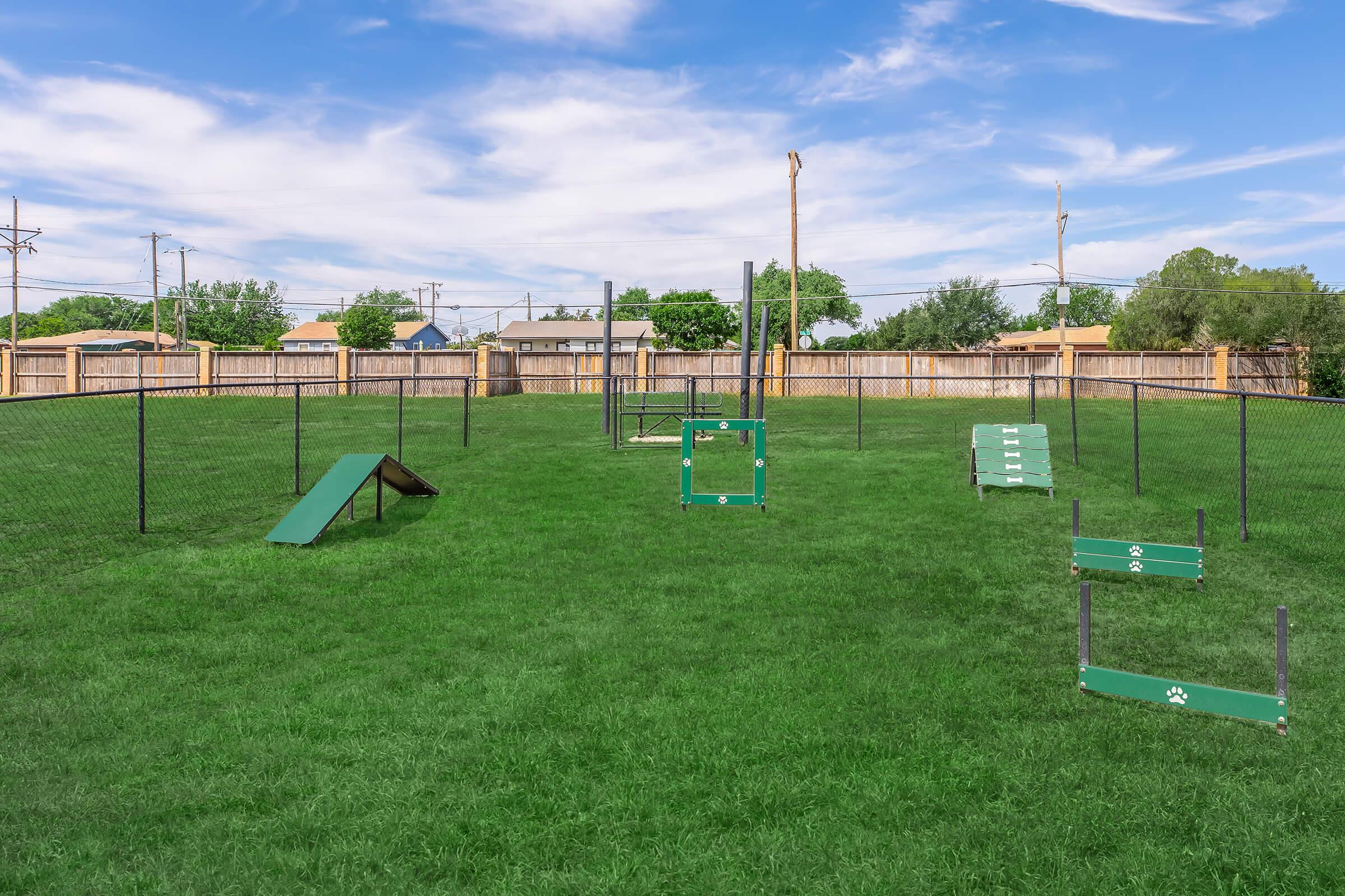 A spacious dog park with green grass enclosed by a black fence. It features various agility equipment, including an A-frame ramp, a small jump, and a frame structure, all in bright green. The sky is clear with fluffy white clouds, and there are trees and houses visible in the background.