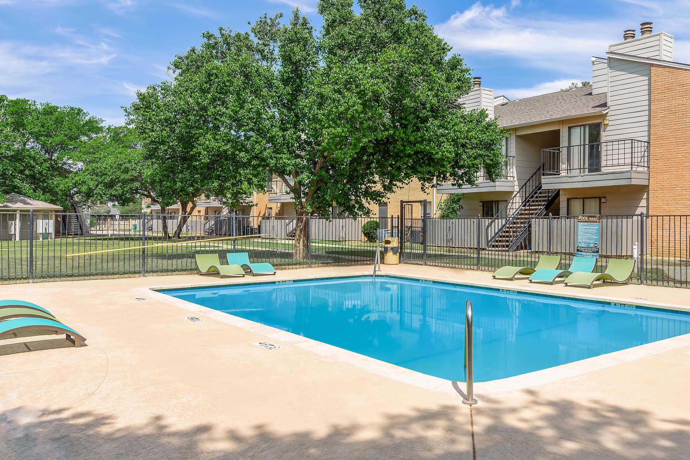 A clear blue swimming pool surrounded by lounge chairs, set in a residential area with green trees and apartment buildings in the background. The area is well-maintained, with a fenced perimeter, and a bright blue sky above.