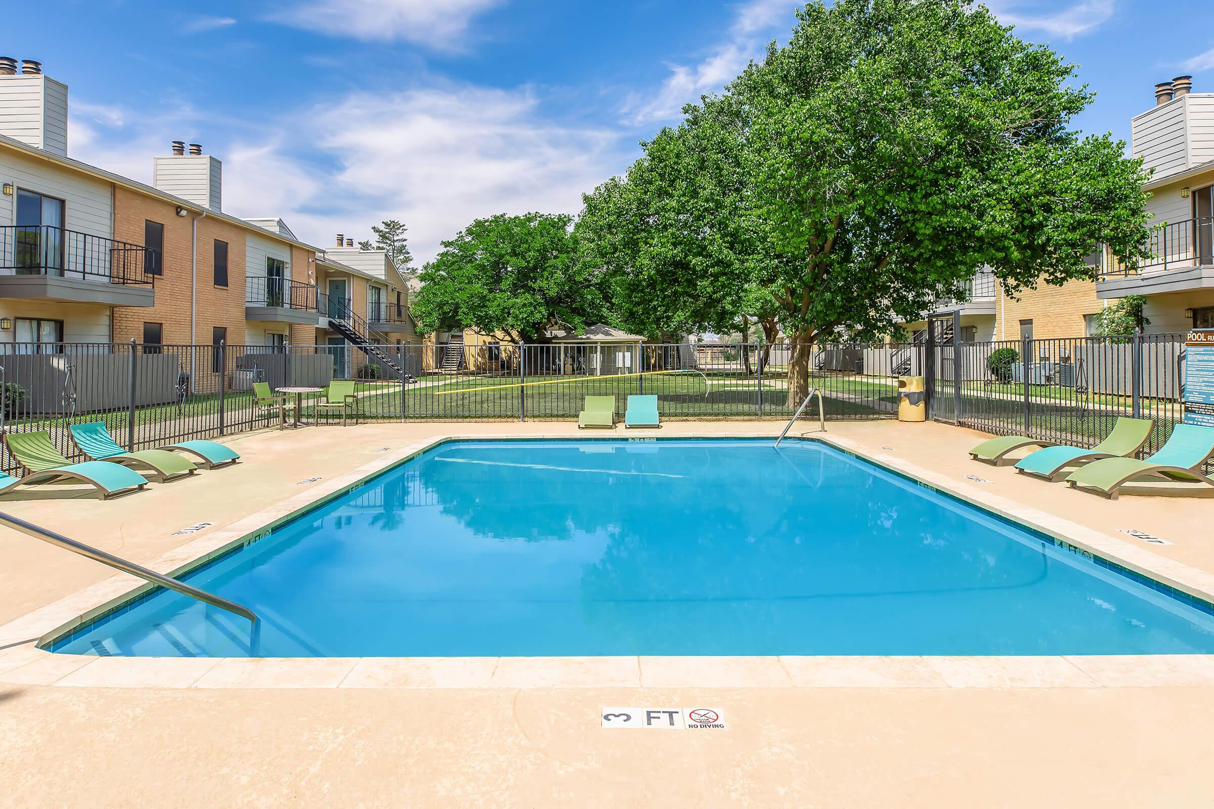 A serene outdoor swimming pool surrounded by lounge chairs and a grassy area, with apartment buildings in the background under a blue sky. The pool has a clear, inviting water surface and a shallow end marked with a depth of 3 feet.