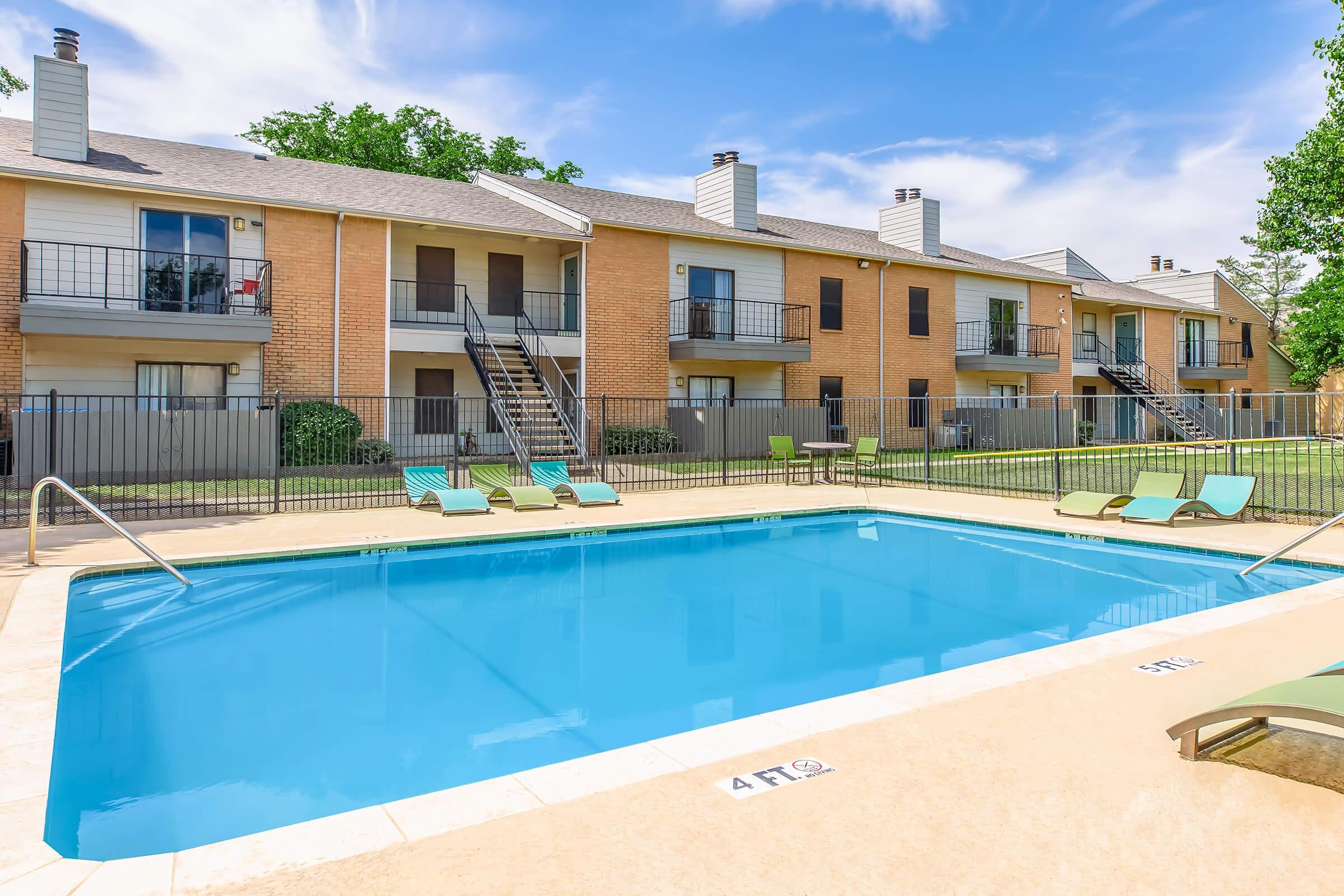 A clear blue swimming pool surrounded by green lounge chairs, with two-story apartment buildings in the background. The pool area is enclosed by a fence, and the sky is bright with a few clouds. The setting is calm and inviting, ideal for relaxation.