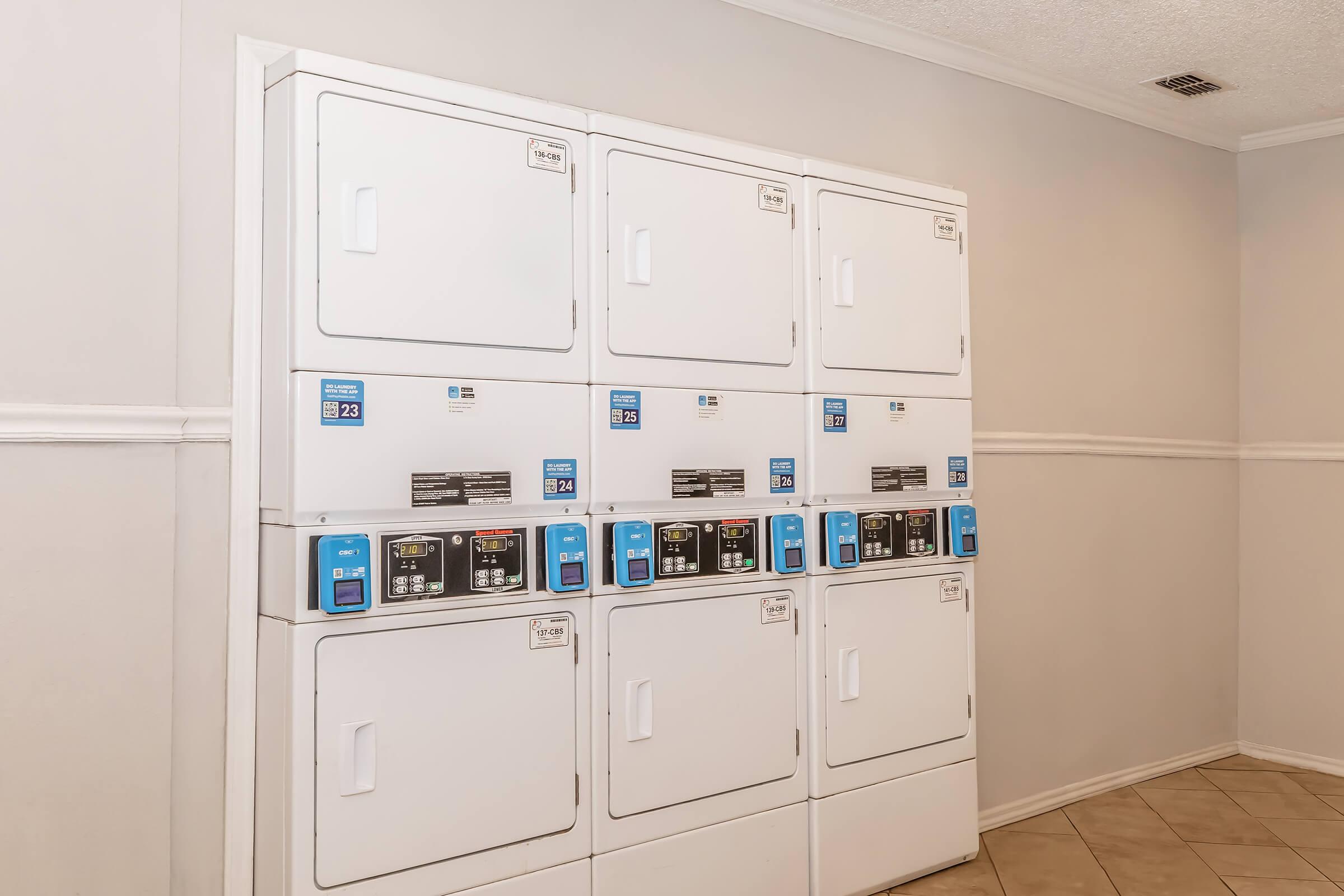 A row of white laundry machines stacked against a light-colored wall, featuring several digital control panels for selecting wash and dry settings. The flooring has beige tiles, and the space appears clean and organized, suitable for laundry activities.