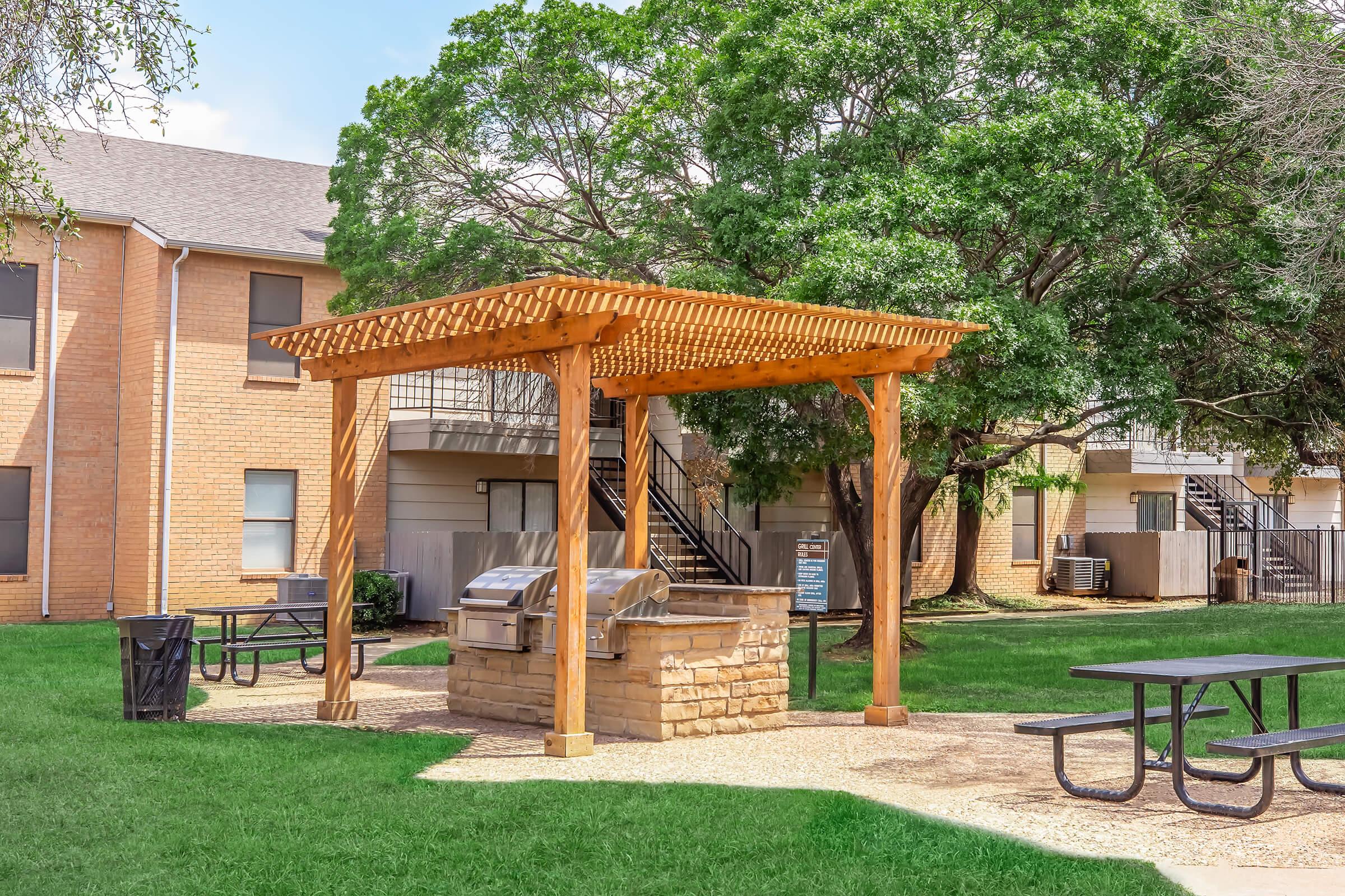 An outdoor grilling area featuring a wooden pergola, stone grill, and picnic tables on a grassy lawn. Surrounded by trees and apartment buildings, it provides a communal space for outdoor cooking and dining. Trash can is visible nearby. Clear blue sky above adds to the inviting atmosphere.