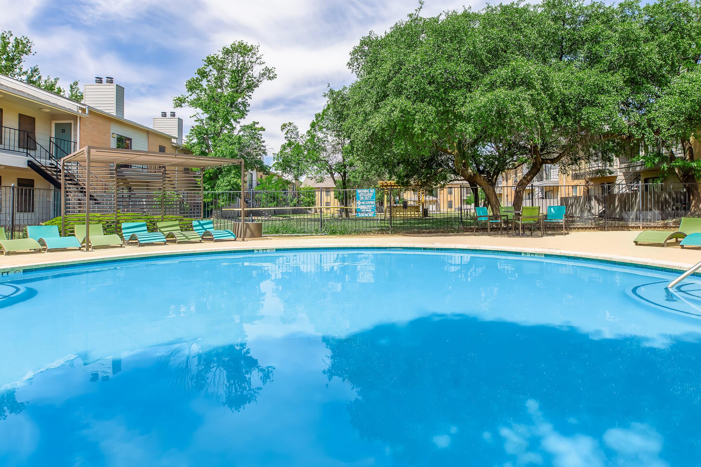 A sparkling blue swimming pool surrounded by green trees and lounge chairs, with a wooden trellis in the background. The pool reflects the clear sky and nearby structures, creating a serene and inviting atmosphere. Perfect for relaxation and outdoor enjoyment.