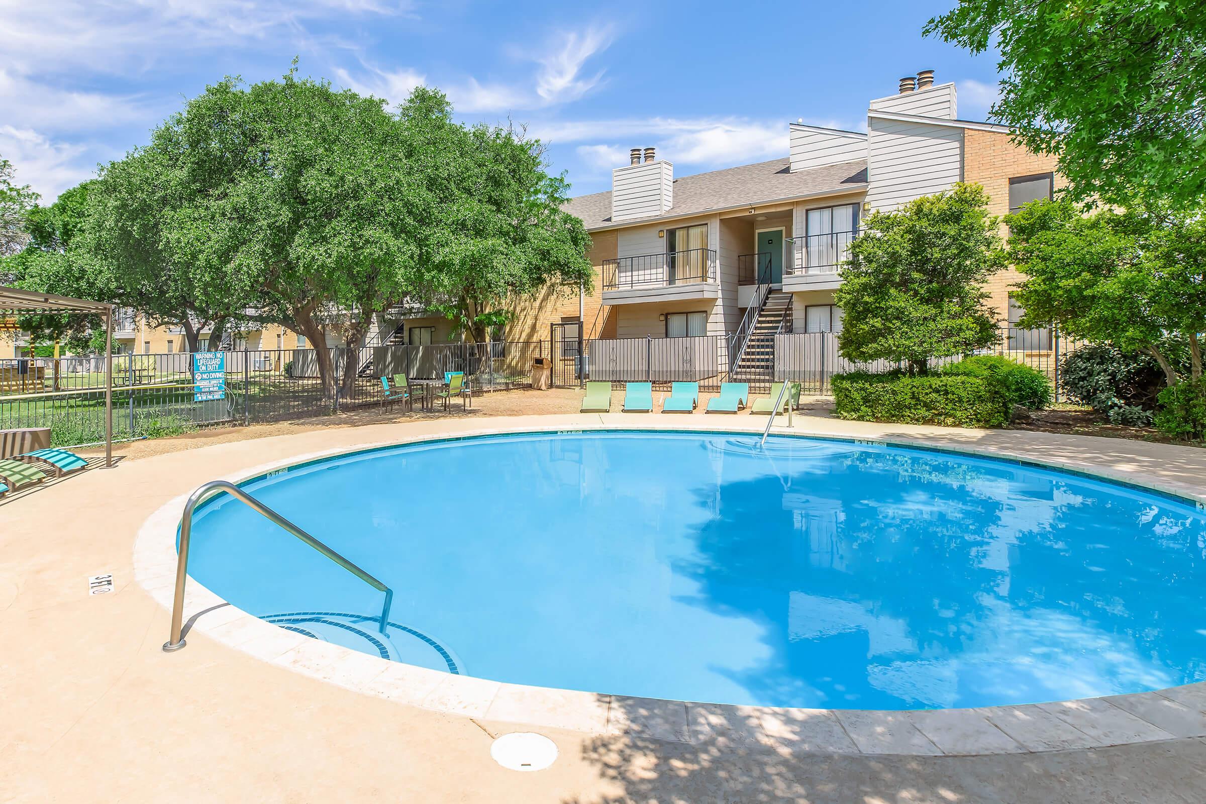 A clear blue swimming pool surrounded by a concrete deck, with poolside lounge chairs and lush green trees nearby. Two apartment buildings are visible in the background, featuring balconies and outdoor areas. The sky is bright with scattered clouds, creating a sunny, inviting atmosphere.