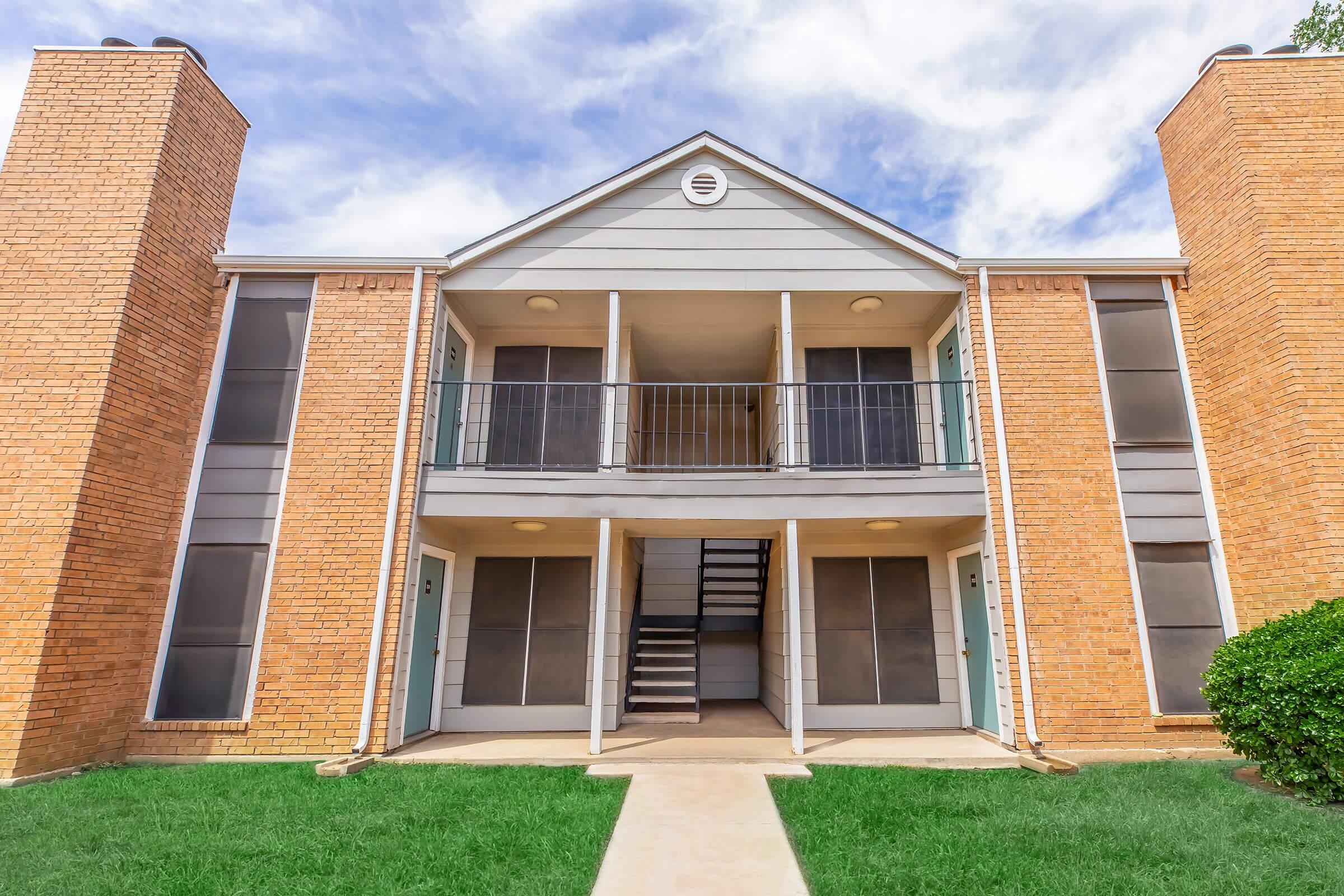 A two-story apartment building with a brick exterior and light gray siding. The entrance features a central staircase leading to the upper floor, with balconies on both levels. Large windows are flanked by two brick chimneys. The surrounding area has well-maintained lawn grass and a pathway leading to the entrance.