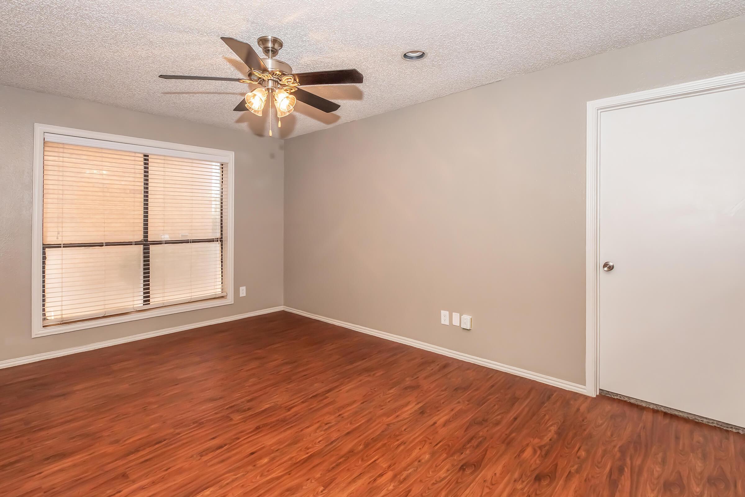 Empty room with light brown wooden flooring, a ceiling fan with four lights, and a window with horizontal blinds. The walls are a light gray color, and there is a white door on the right side. The overall atmosphere is bright and spacious, suitable for living or office use.