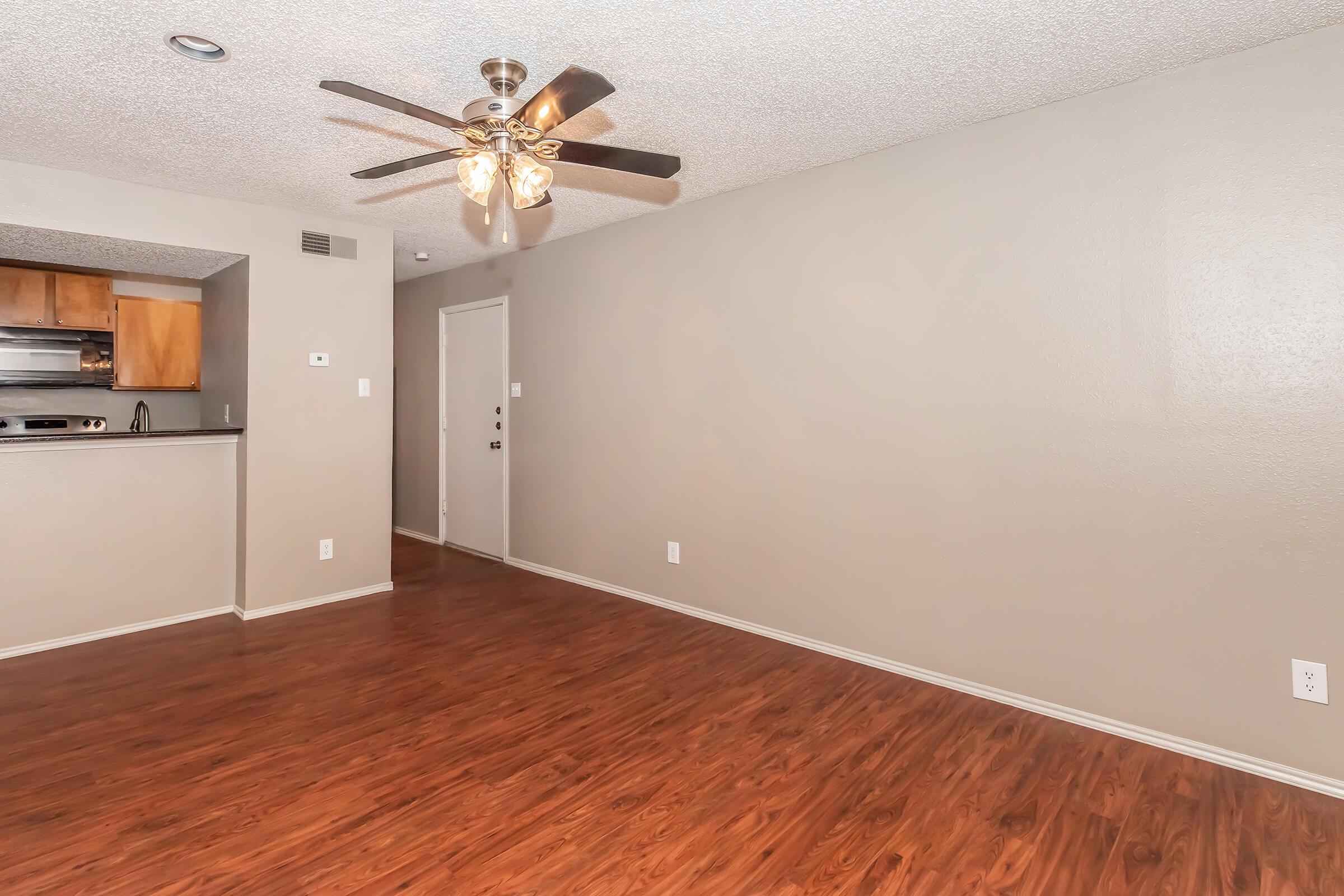 A spacious, empty living room featuring light brown laminate flooring, a ceiling fan with light fixtures, and a pale gray wall. The open layout includes a glimpse of a kitchen area with wooden cabinetry in the background. A doorway leads to an entrance.