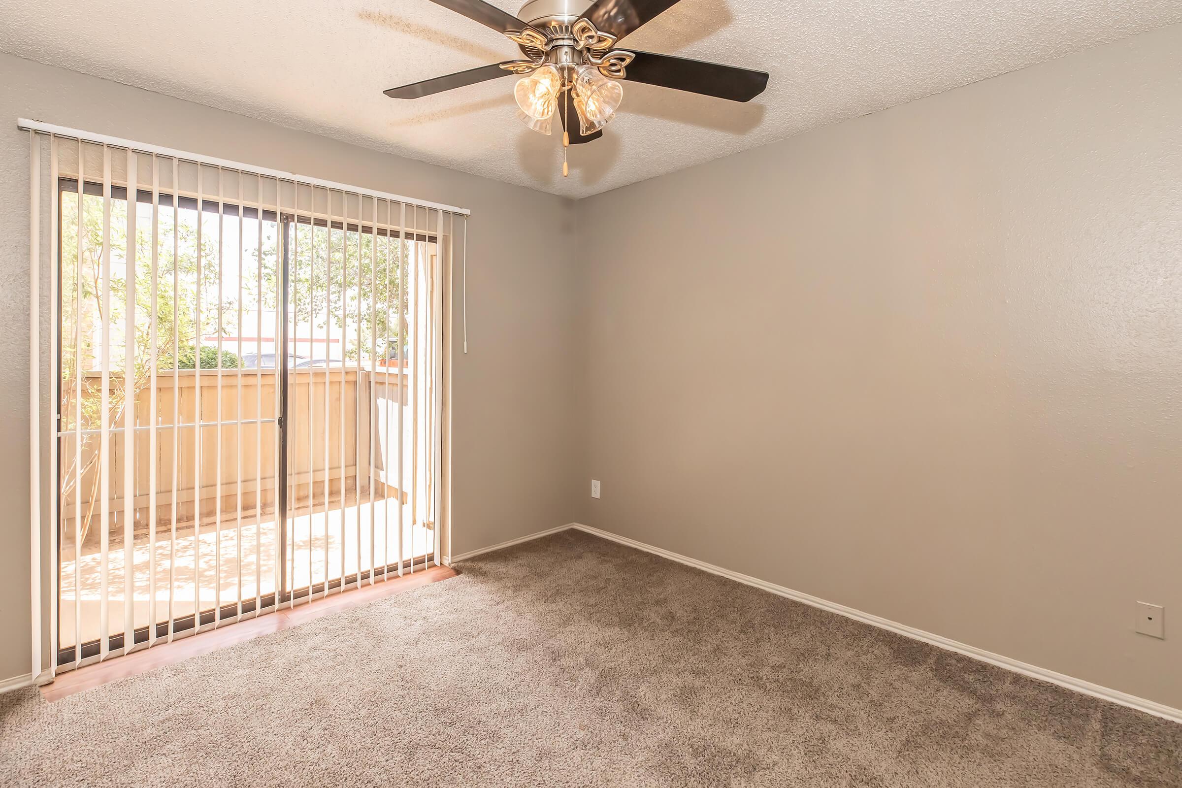 A spacious, empty room featuring light gray walls and a plush carpet. It has a ceiling fan with light fixtures and a large sliding glass door with vertical blinds that leads to a small balcony area. Natural light illuminates the space, creating a bright and inviting atmosphere.