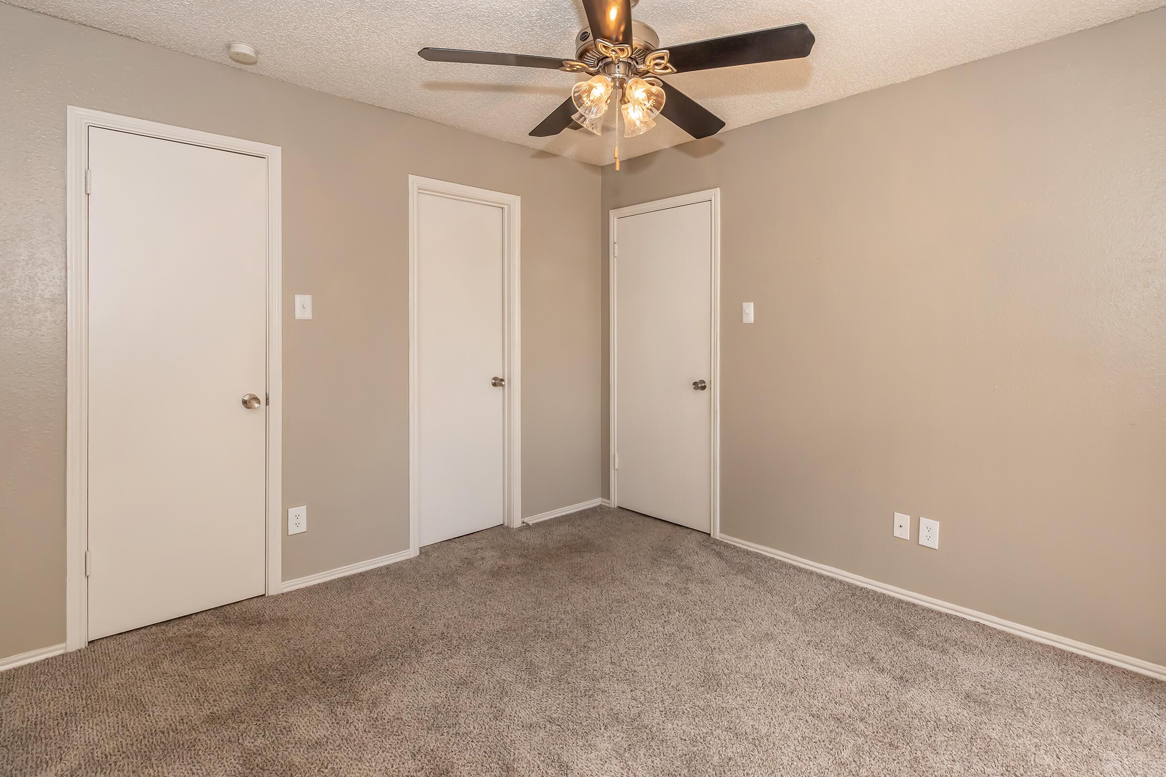 A neat, empty room featuring gray walls and light gray carpet. There are three closed white doors on two sides and a ceiling fan with light fixtures in the center. The space has a clean and minimalistic appearance, ideal for a bedroom or office setup.