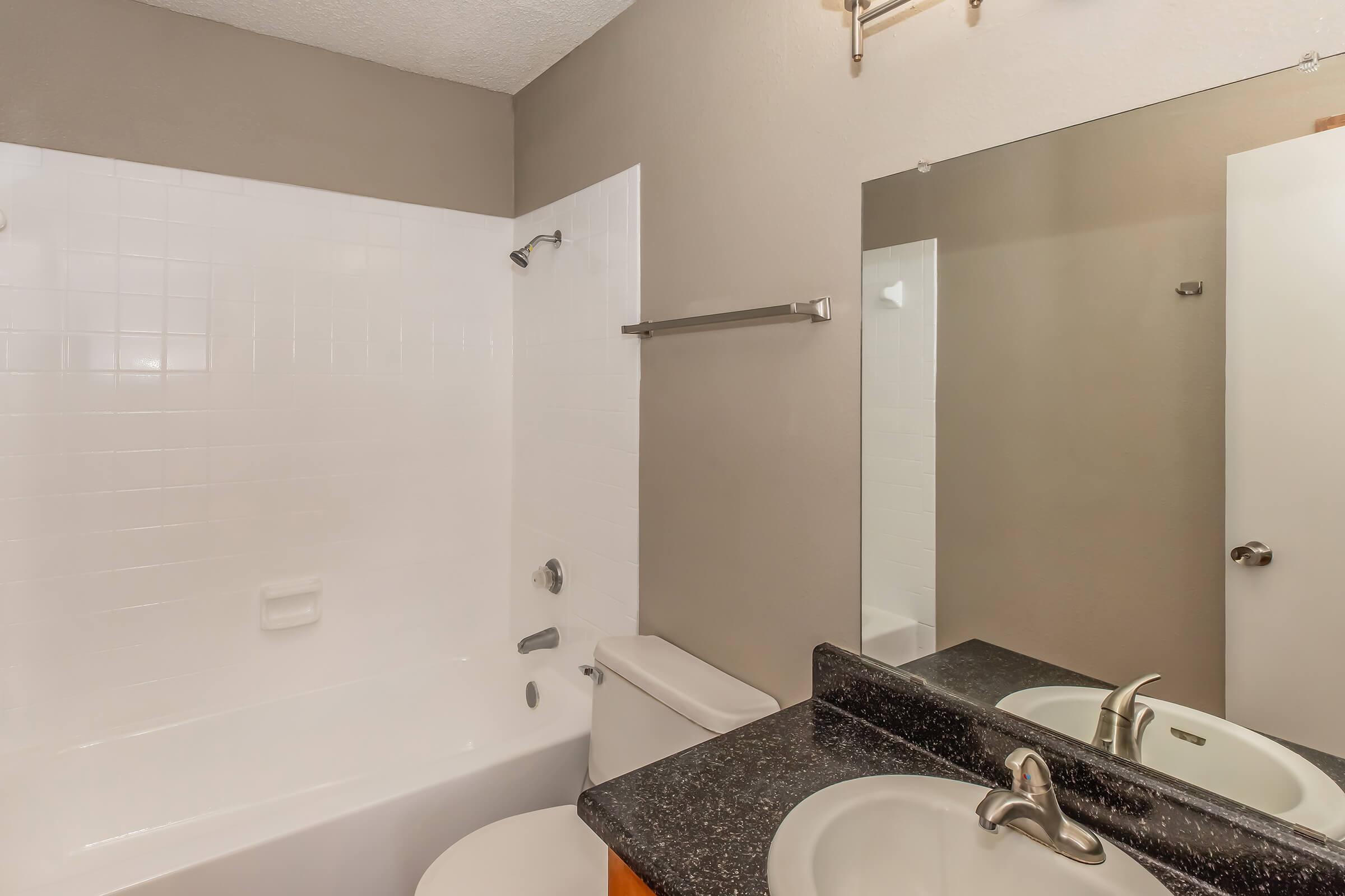 A modern bathroom featuring a white bathtub with a shower, beige walls, a black countertop with a sink, and a large mirror. There's a towel rack and a toilet visible, creating a clean and functional space.