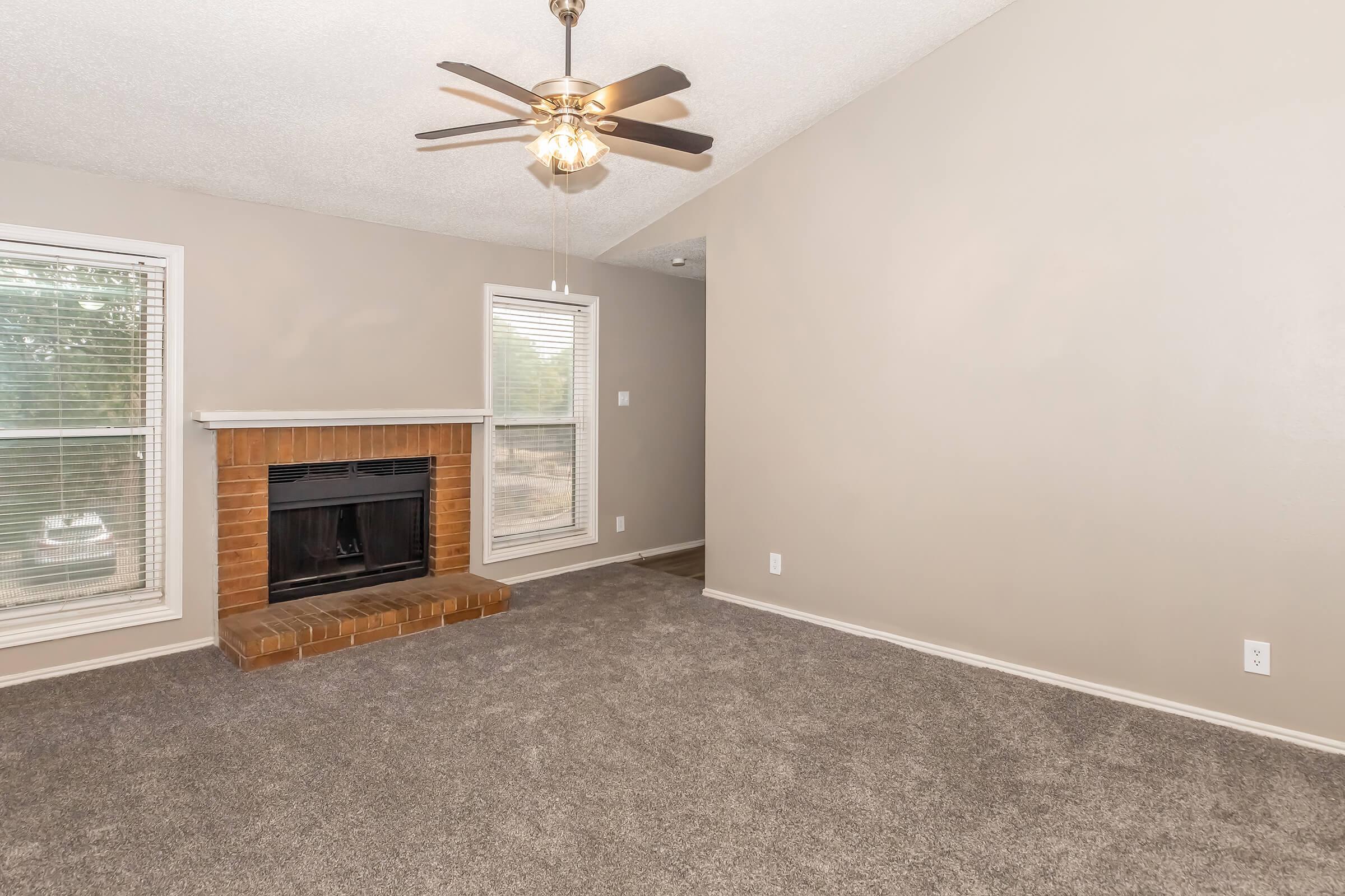 A spacious living room featuring a ceiling fan, a brick fireplace, and large windows allowing natural light. The walls are painted a neutral color, and the floor is covered with gray carpet. There is a visible transition to another room, adding to the open feel of the space.