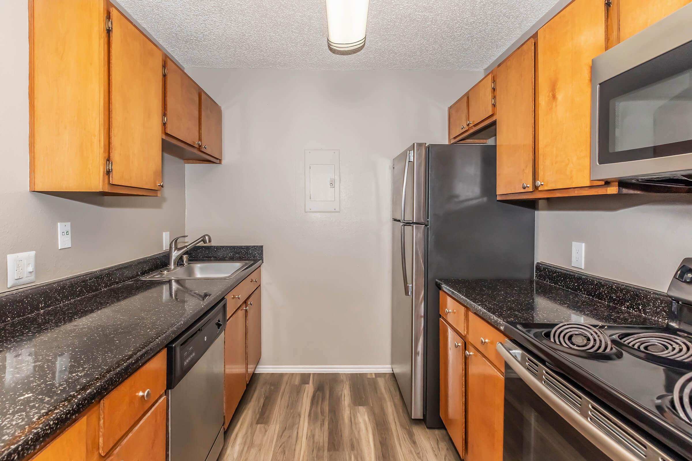 A modern kitchen featuring wooden cabinetry, black granite countertops, and stainless steel appliances including a refrigerator, microwave, and stove. The flooring is faux wood, and the space is well-lit with overhead lighting, creating a clean and contemporary atmosphere.