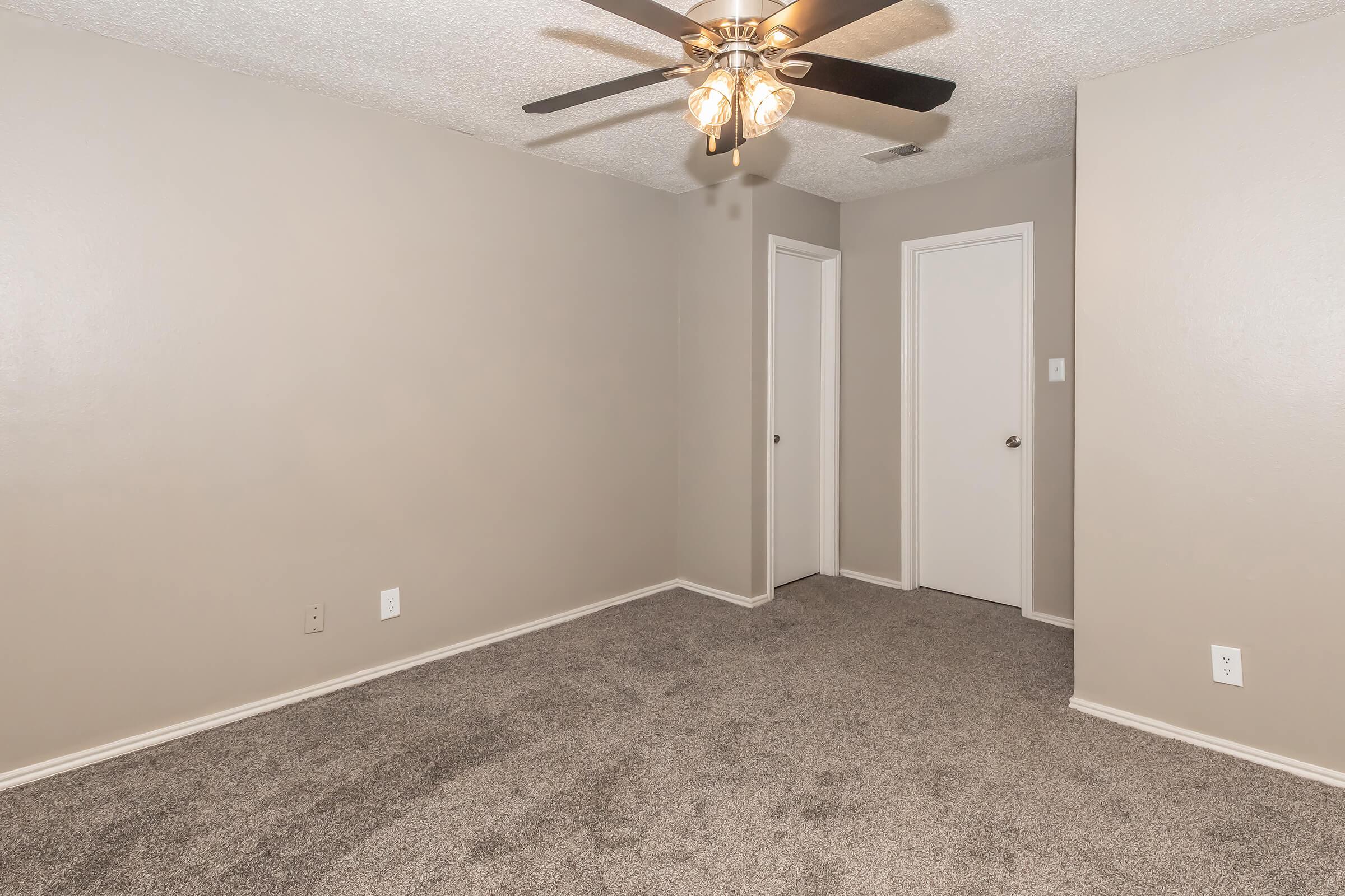 An interior view of a small, empty room featuring light beige walls and plush gray carpet. A ceiling fan with five light bulbs hangs from the ceiling, and there are two closed white doors on one wall, adding to the minimalist aesthetic of the space.