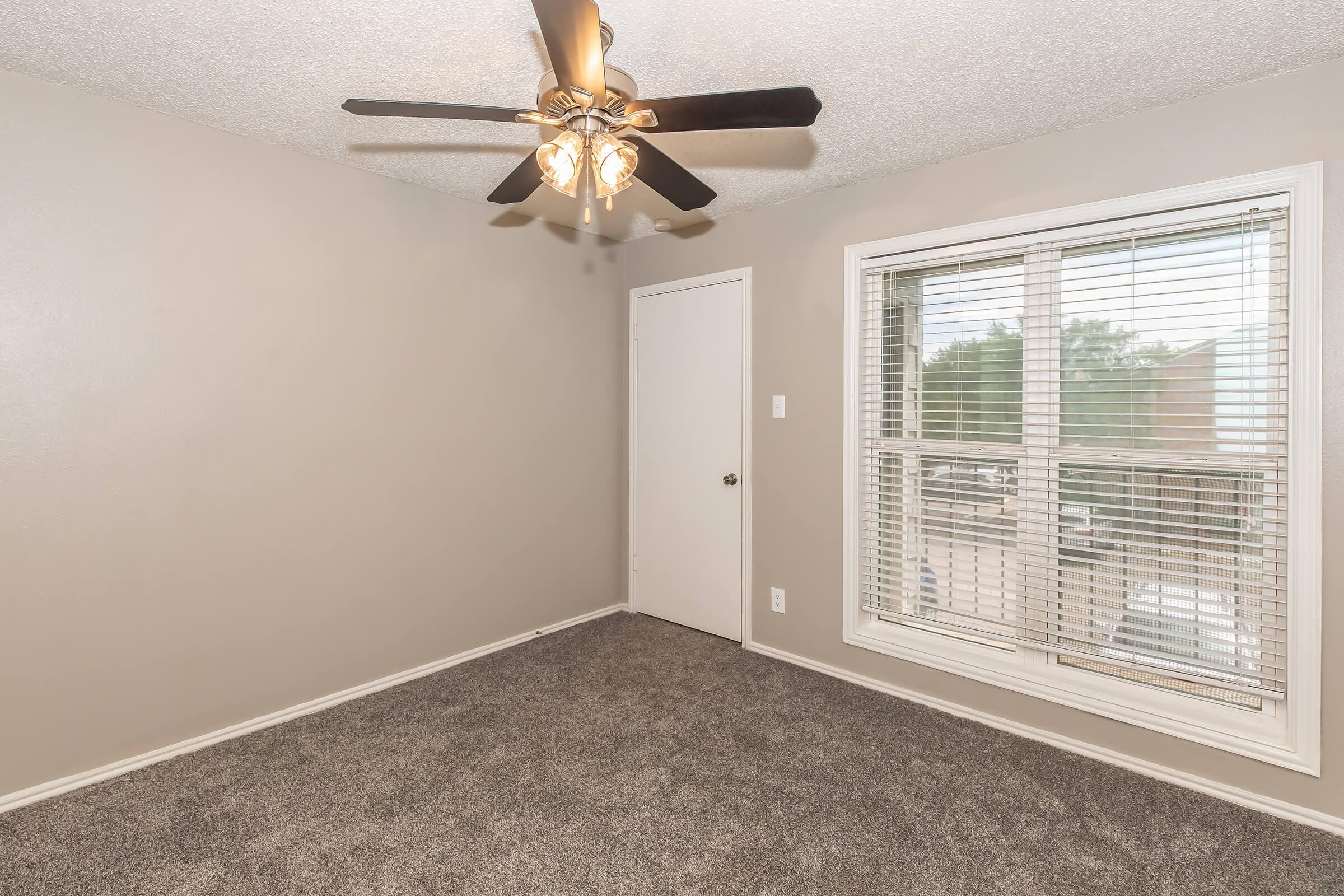 A well-lit room with gray walls and carpet, featuring a ceiling fan with three lights. A closed door is visible on one wall, and a large window with white blinds lets in natural light, overlooking an outdoor area. The overall ambiance is cozy and inviting.